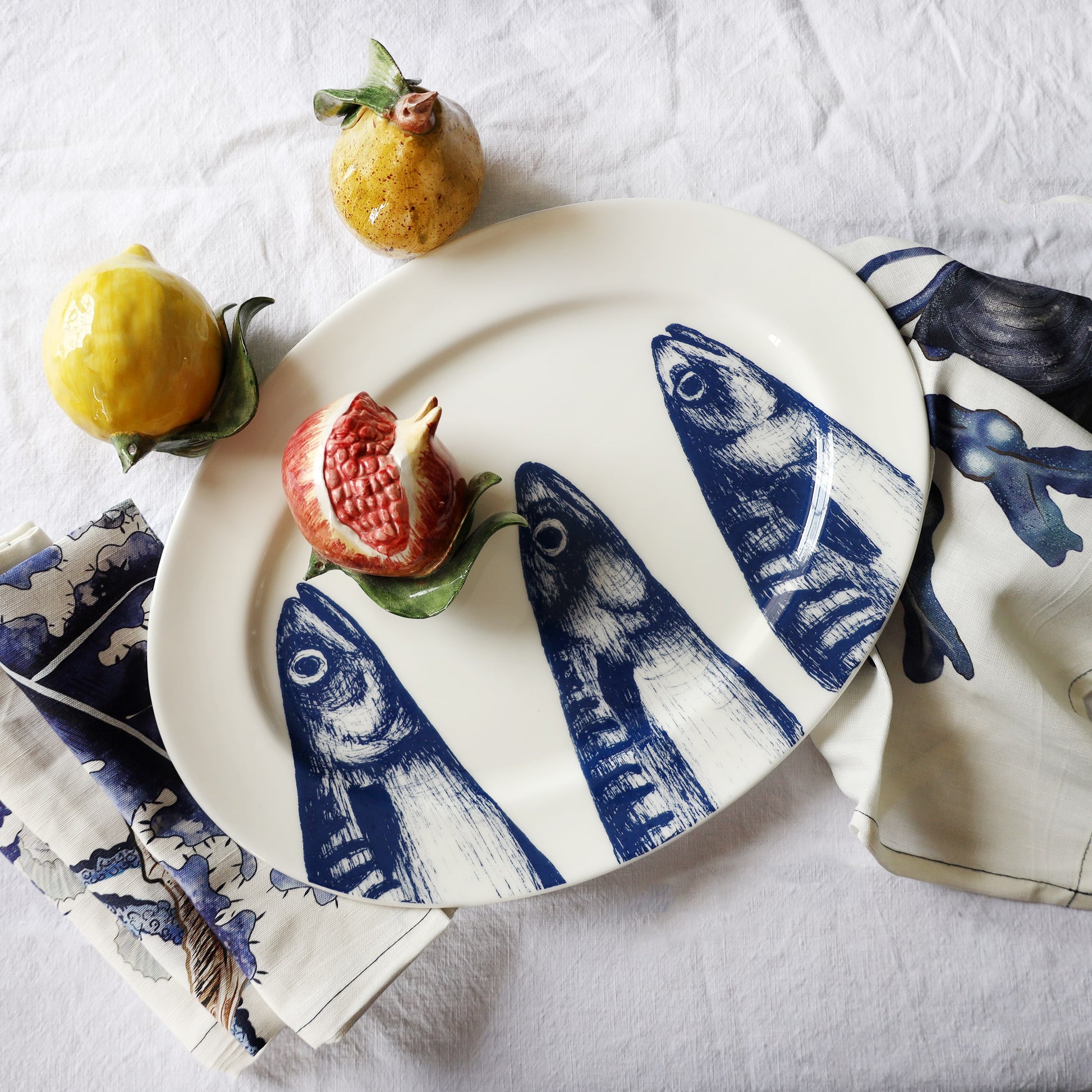 White plate with blue mackerel heads design, accompanied by lemons and pomegranate on a white surface.