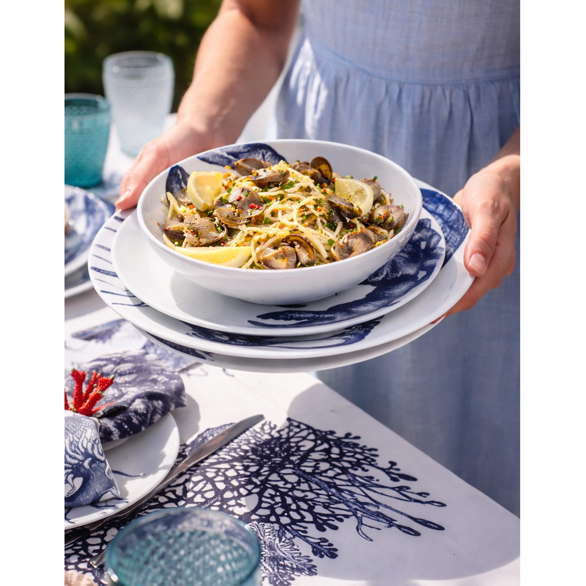 Person holding a bowl of pasta with a blue and white patterned tablecloth in the background