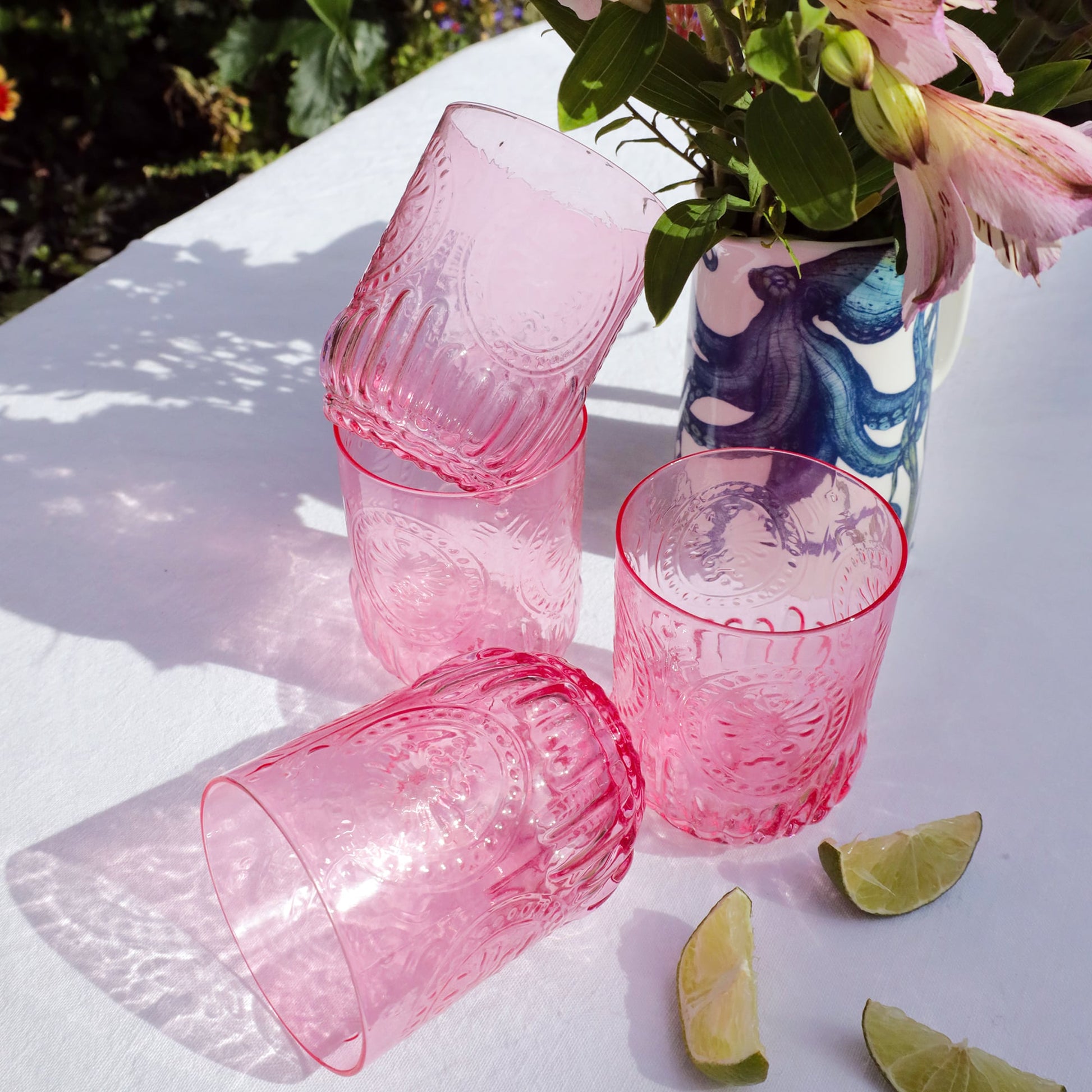 Stack of pink textured glasses on a white surface with flowers and lime wedges.