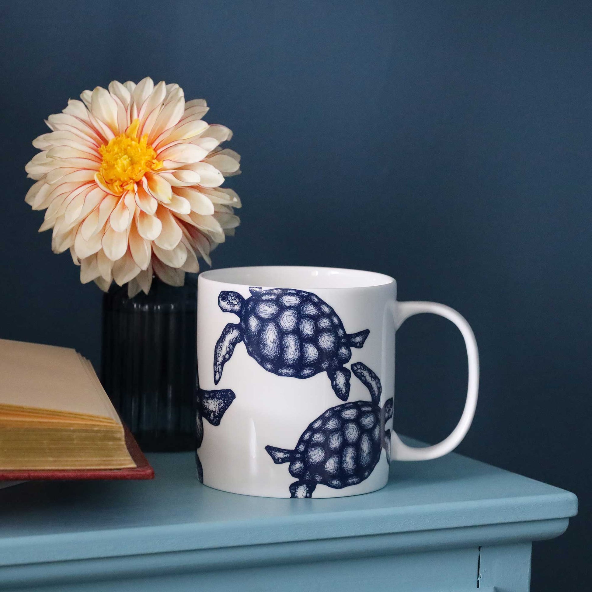 A white ceramic mug with a navy blue turtle pattern, styled on a light blue table next to a black vase with a cream dahlia flower and an open book against a dark blue background.