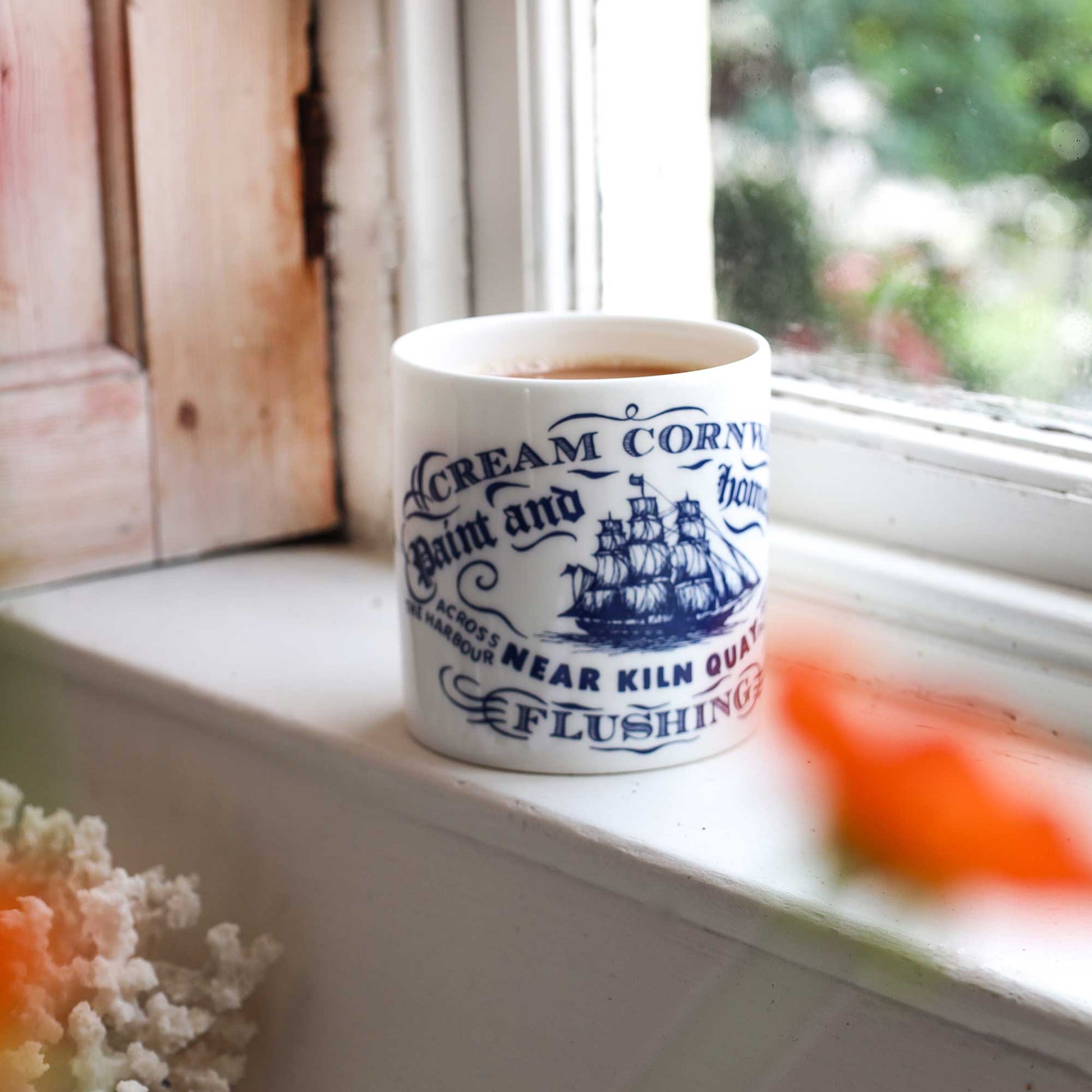 A white ceramic mug with blue printed text and a ship illustration sits on a window sill. The mug reads 'Cream Cornish Paint and Homewares Near Kiln  Flushing'.