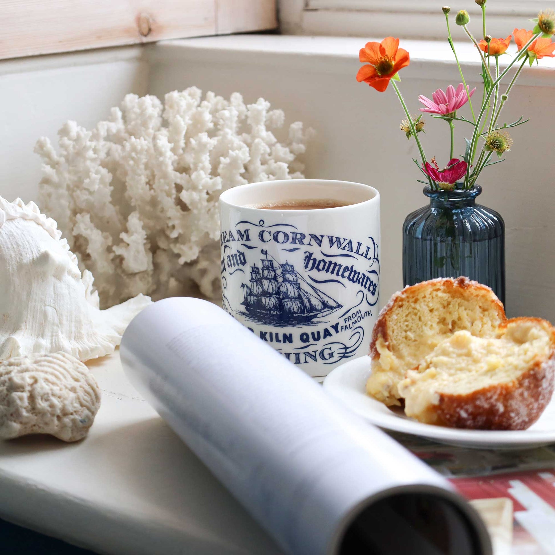 A white ceramic mug with blue printed text and ship illustrations sits on a windowsill beside a plate with a sliced pastry, a rolled paper tube, seashells, coral, and a small blue vase with orange and pink flowers.