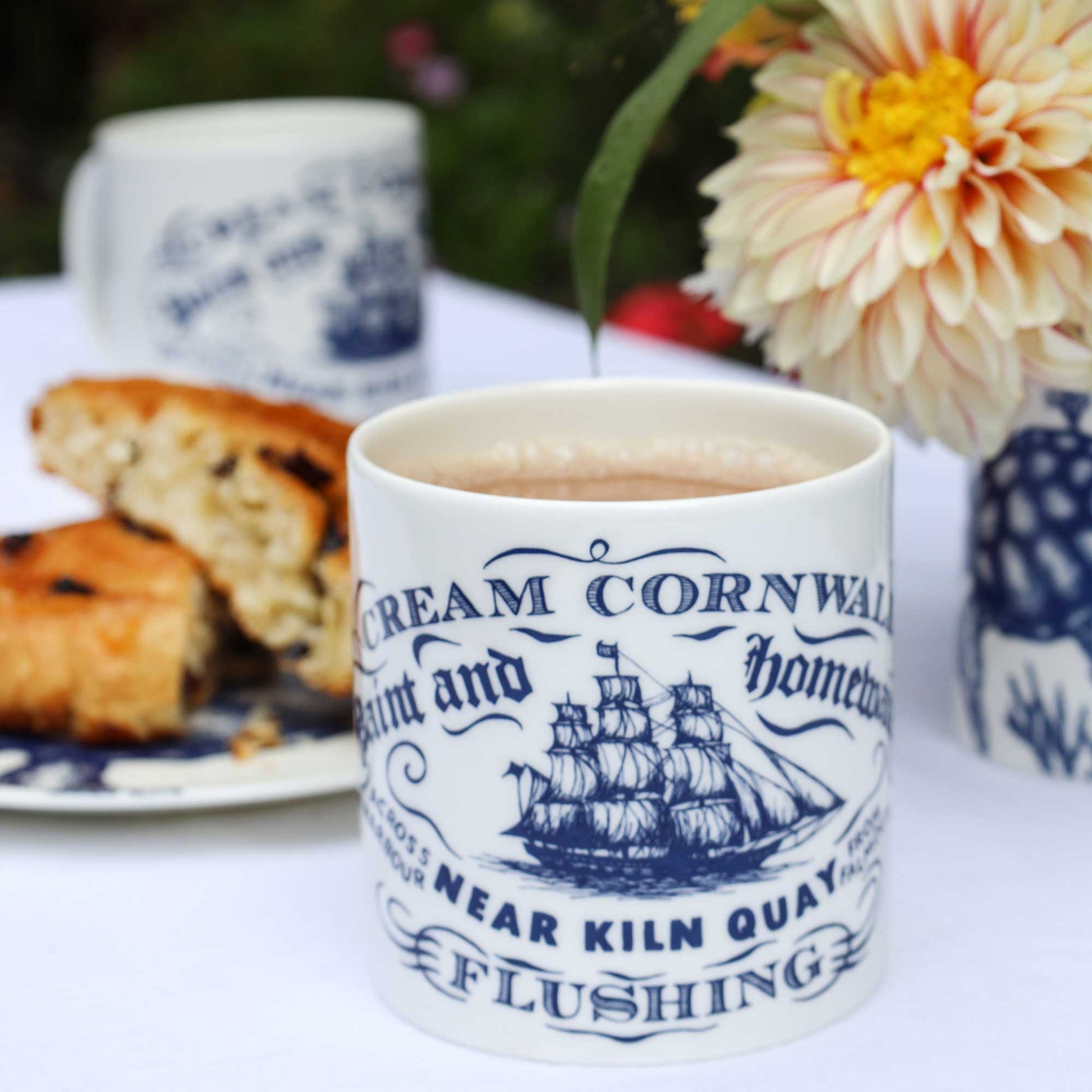 A white ceramic mug with blue printed text reading 'Cream Cornwall' and a navy ship illustration, filled with tea and placed on a table with scones, another mug, and flowers in the background.