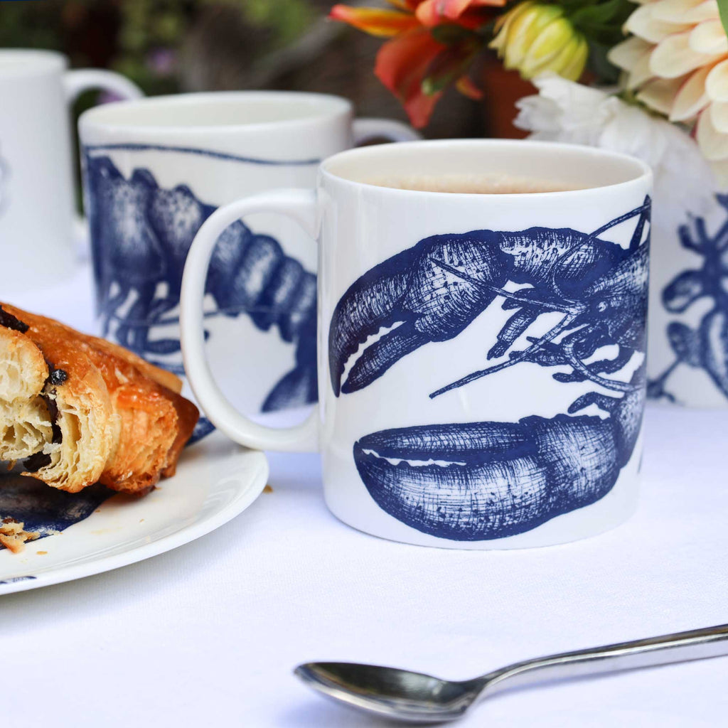 A white ceramic mug with a navy blue lobster illustration, filled with coffee and set on a white tablecloth next to a plate with a pastry and a spoon. Another matching mug and a floral arrangement are blurred in the background.