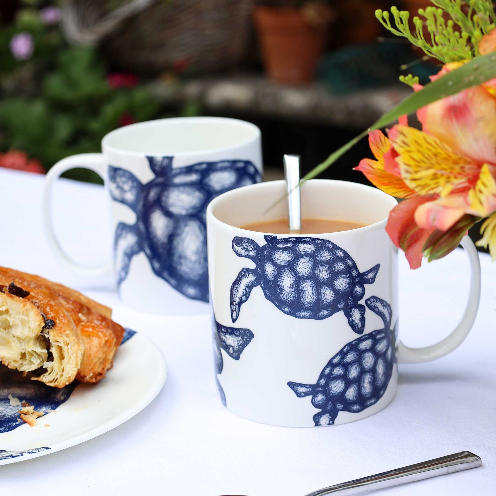 Two white ceramic mugs with dark blue turtle illustrations, one filled with tea and one empty, set on a white tablecloth with a plate of pastry and flowers in the background.