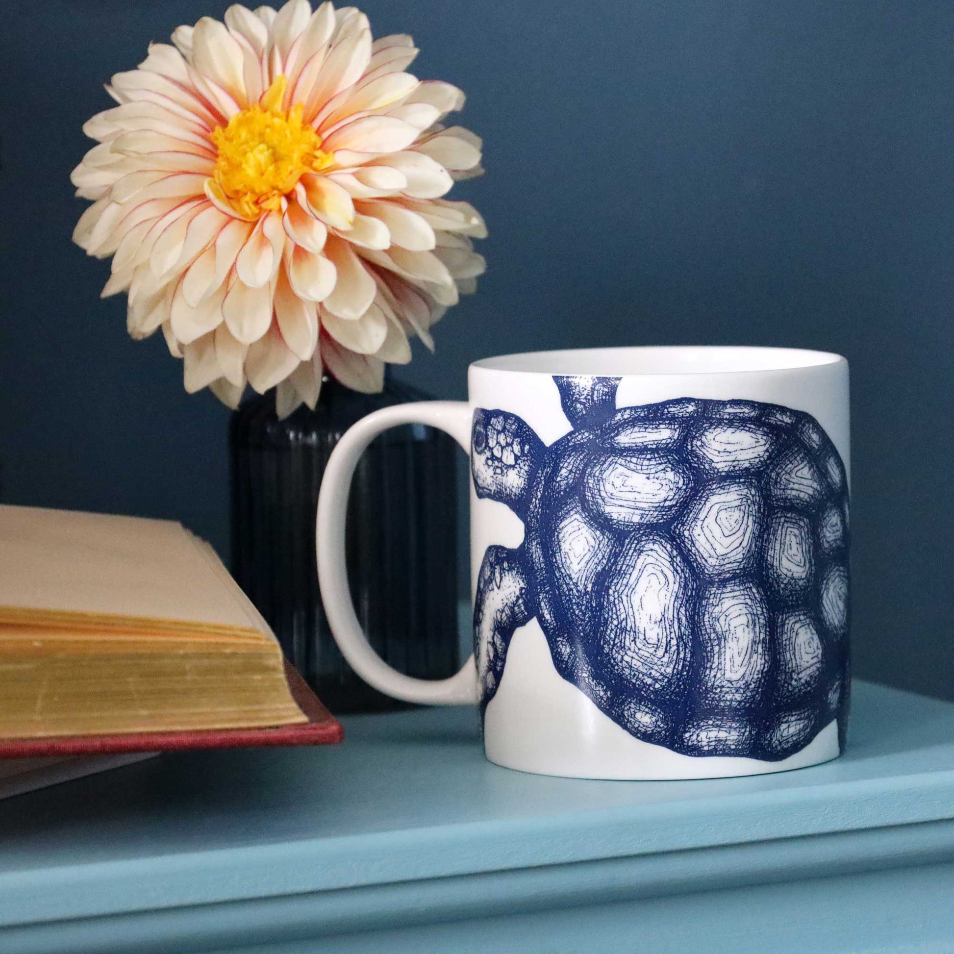 A white ceramic mug with a navy blue turtle illustration, styled on a light blue shelf next to a book and a flower in a vase against a dark blue background.