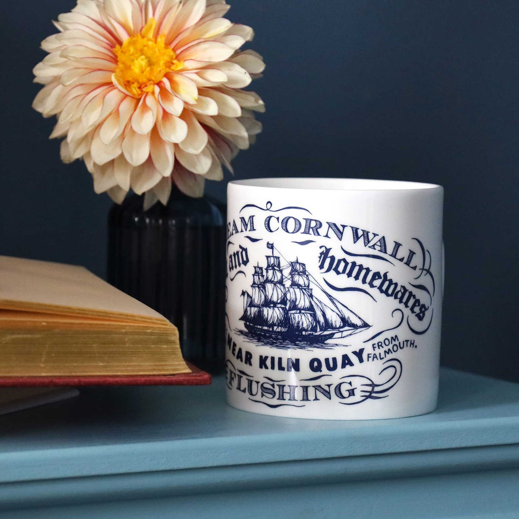 A white ceramic mug with a navy blue printed design of a sailing ship and the text 'Cream Cornwall and Homewares Near Kiln Quay from Falmouth Flushing,' styled on a blue surface next to a book and a flower in a dark vase against a dark blue background.