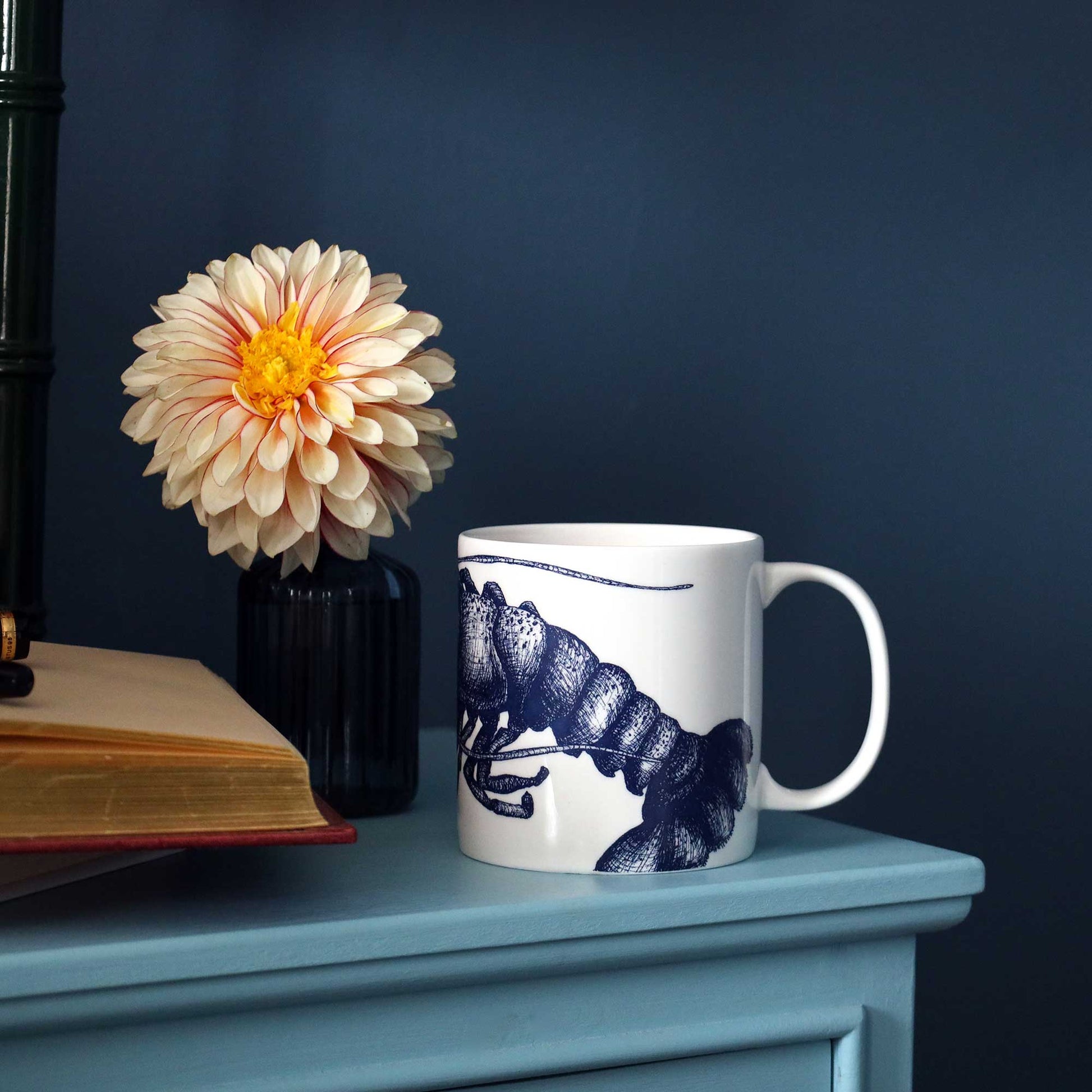 A white ceramic mug with a navy blue lobster illustration sits on a dusty blue side table next to a black vase holding a cream and orange dahlia flower, with an open book in the background against a dark navy wall.