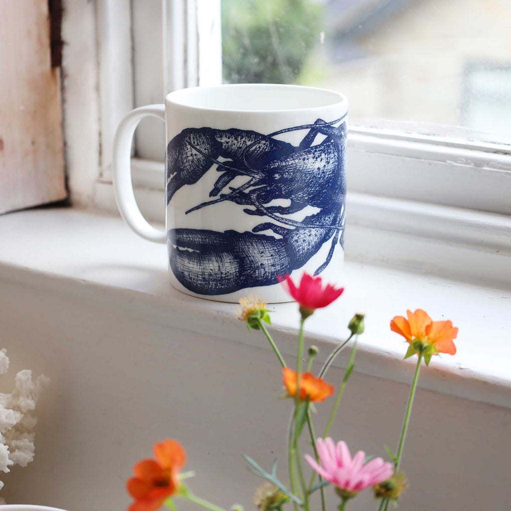 A white ceramic mug with a dark blue lobster illustration sits on a white windowsill, with blurred orange and pink flowers in the foreground and a window behind it.
