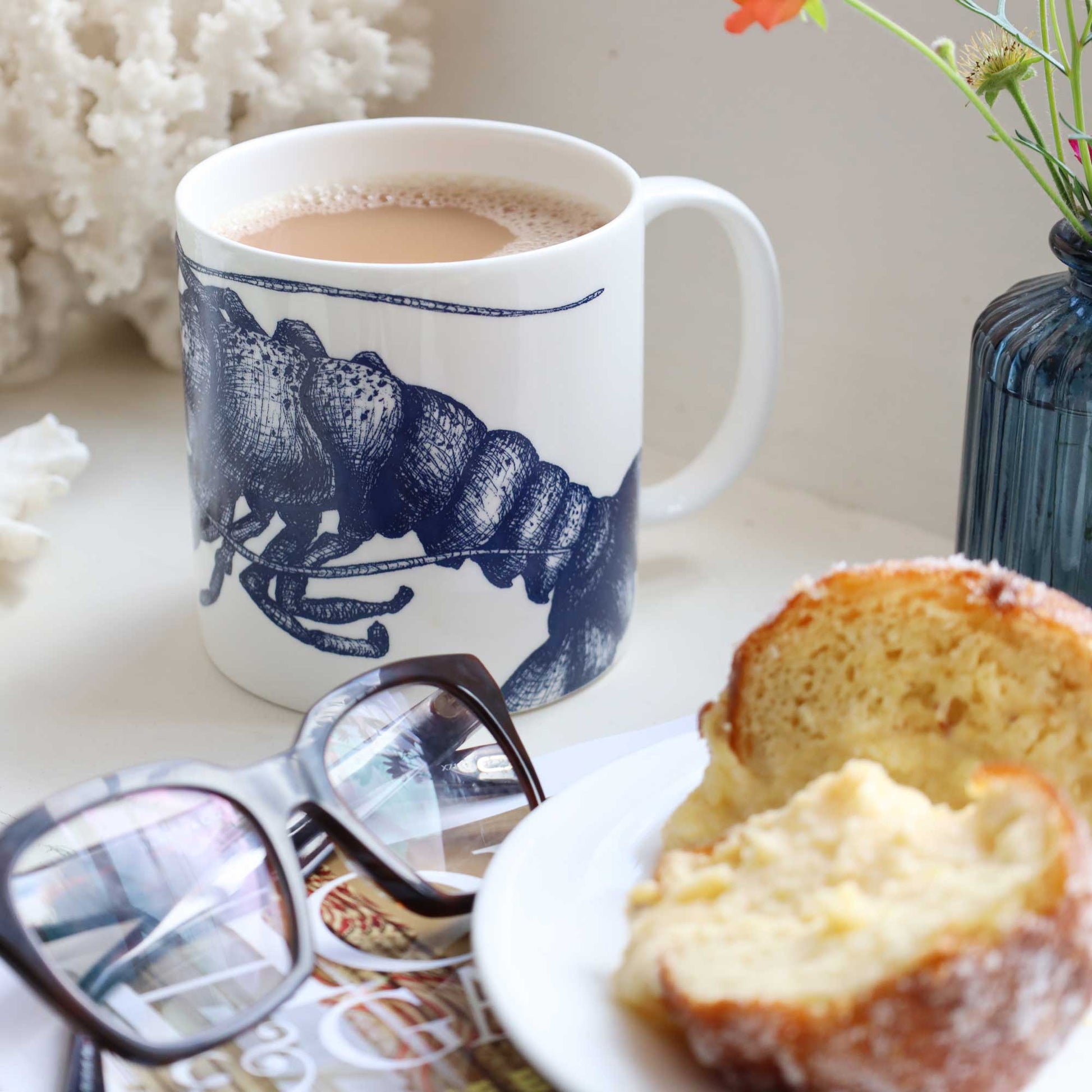A white ceramic mug with a dark blue lobster illustration, filled with a light brown beverage, styled on a tabletop with a slice of toast on a plate, eyeglasses, and a vase in the background.