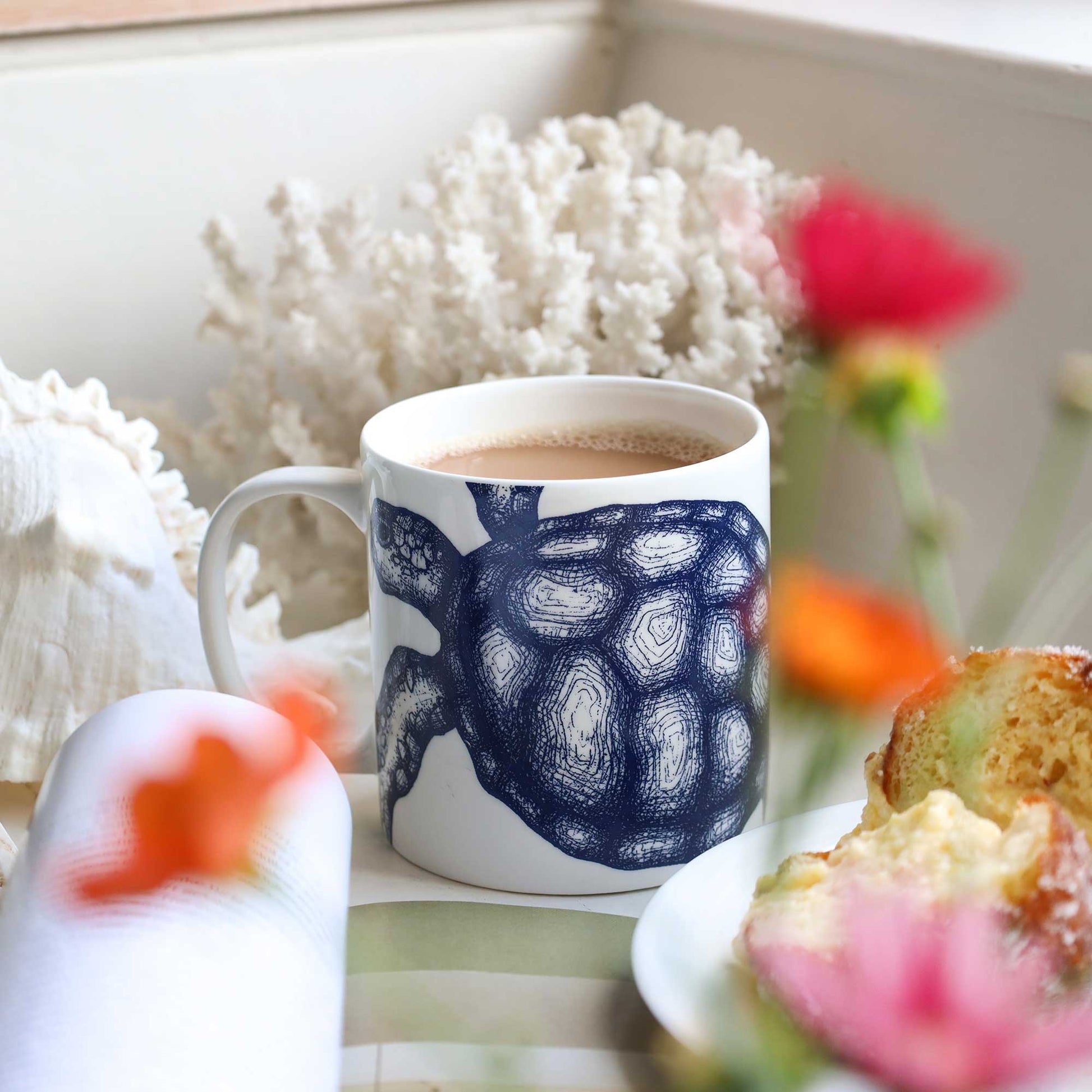 A white ceramic mug with a navy blue turtle illustration, filled with a light brown beverage, styled on a table with a plate of food and blurred flowers in the background.