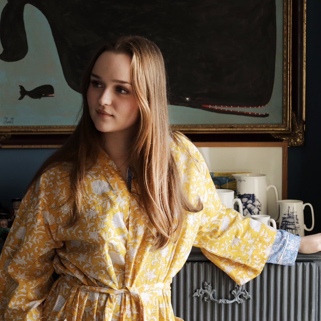 Young woman wearing a yellow block-printed dressing gown with intricate white floral and seashell patterns, standing in a cozy interior. Behind her, a painting of a whale and nautical-themed ceramics add a vintage touch to the decor.