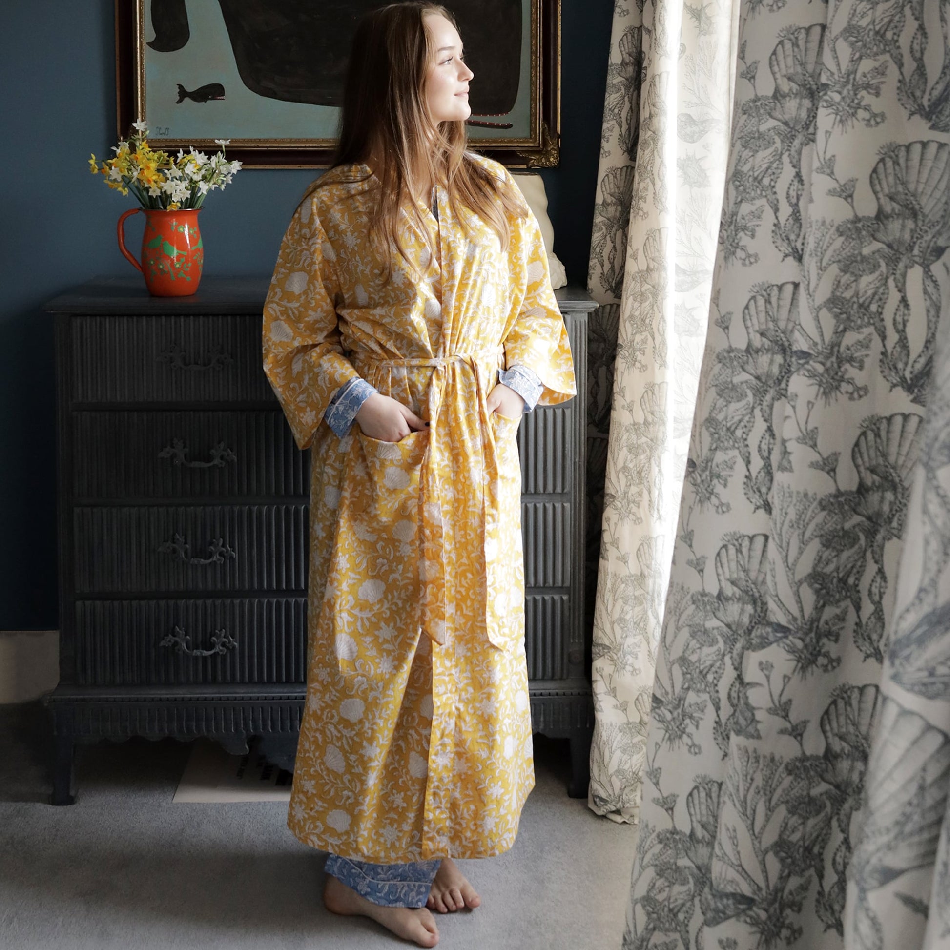 Young woman standing by a window wearing a yellow block-printed dressing gown with white floral and seashell motifs, paired with blue and white patterned pyjama bottoms. The room features a dark grey chest of drawers with a vase of flowers and a framed painting of a whale, while light curtains with intricate botanical patterns frame the scene.