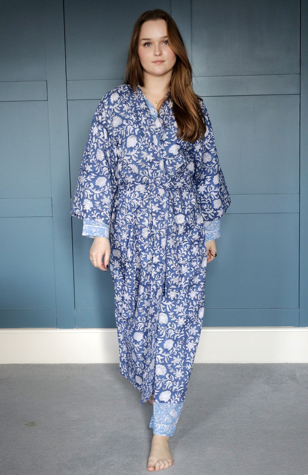 A woman dressed in a long navy blue dressing gown featuring intricate white block-printed floral and shell patterns. The gown has light blue cuffs with matching designs. She stands barefoot on a carpeted floor against a backdrop of panelled blue walls, exuding a calm and elegant appearance.