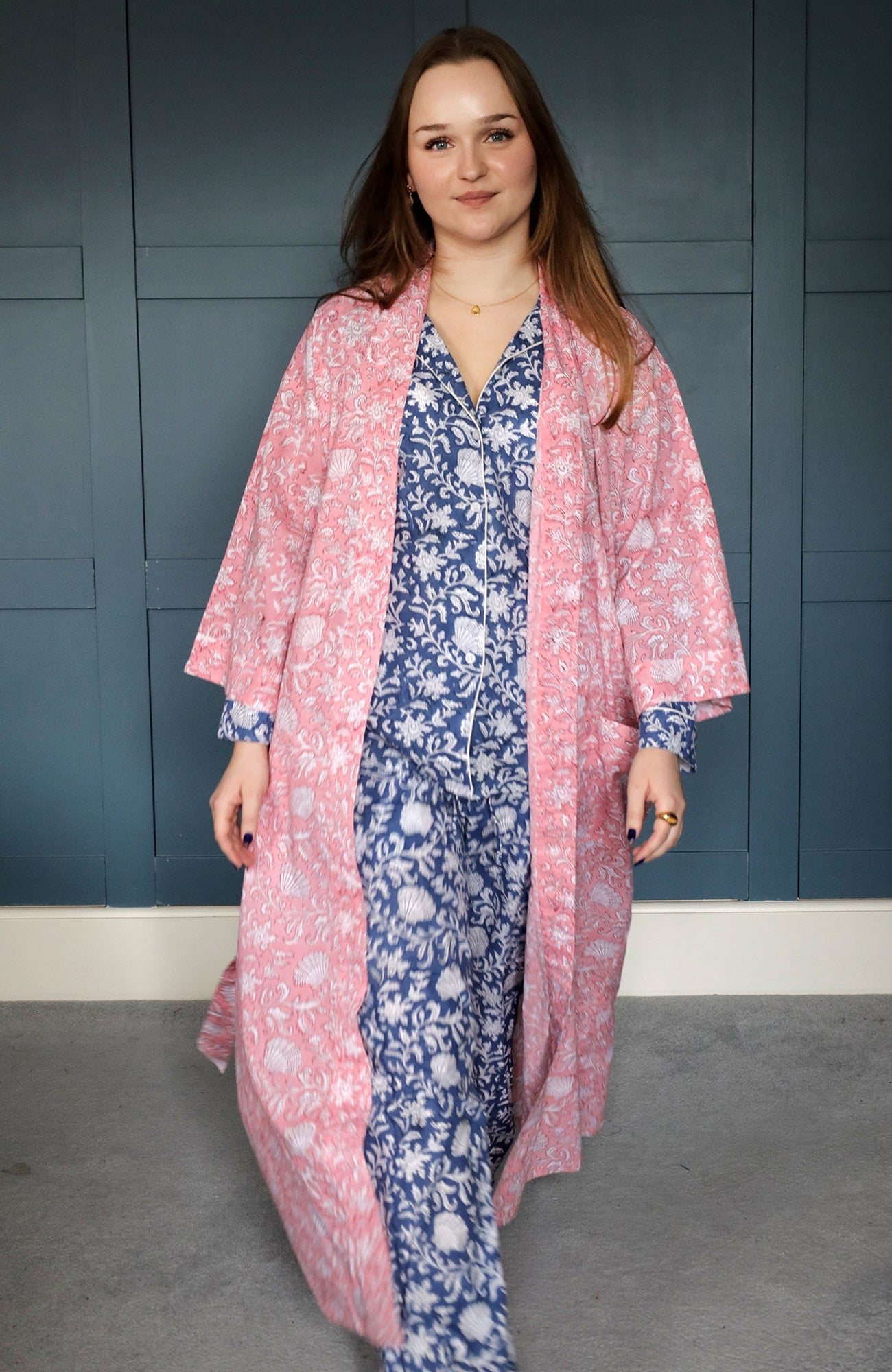 A woman confidently walking indoors wearing a blue block-printed pajama set with white floral and seashell motifs, layered with a flowing pink block-printed dressing gown featuring similar patterns. The background features a dark teal panelled wall, adding a contemporary touch.