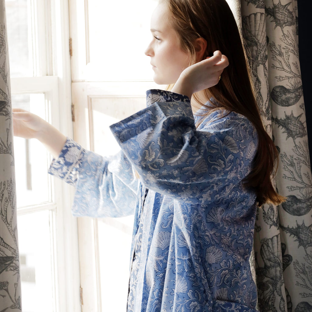 Person standing by a window, gently adjusting the curtain, wearing a light blue dressing gown with intricate white floral and seashell block print patterns. The soft light highlights the delicate design, creating a serene and elegant atmosphere.