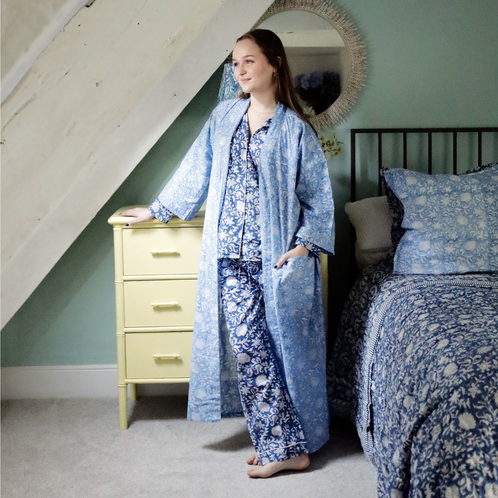 Person standing in a cozy bedroom wearing a light blue dressing gown with intricate white floral and seashell block print patterns, layered over navy floral pyjamas. The room features a yellow dresser, a bed with matching blue and navy block print bedding, and a mirror on a soft green wall.