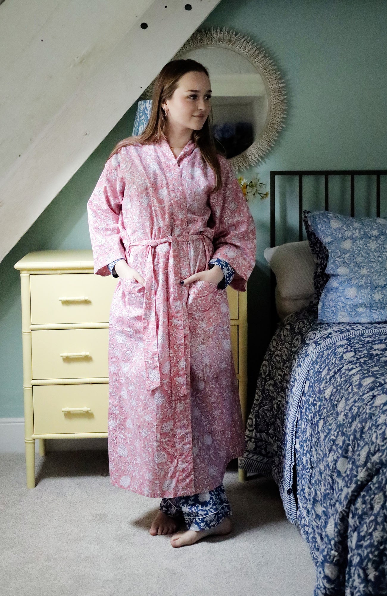 A woman standing in a cozy bedroom wearing a pink block-printed dressing gown with white floral and seashell patterns, tied at the waist. She pairs it with blue block-printed pyjama bottoms. The room features a soft green wall, a yellow chest of drawers, a round decorative mirror, and a bed adorned with blue and white block-printed bedding.