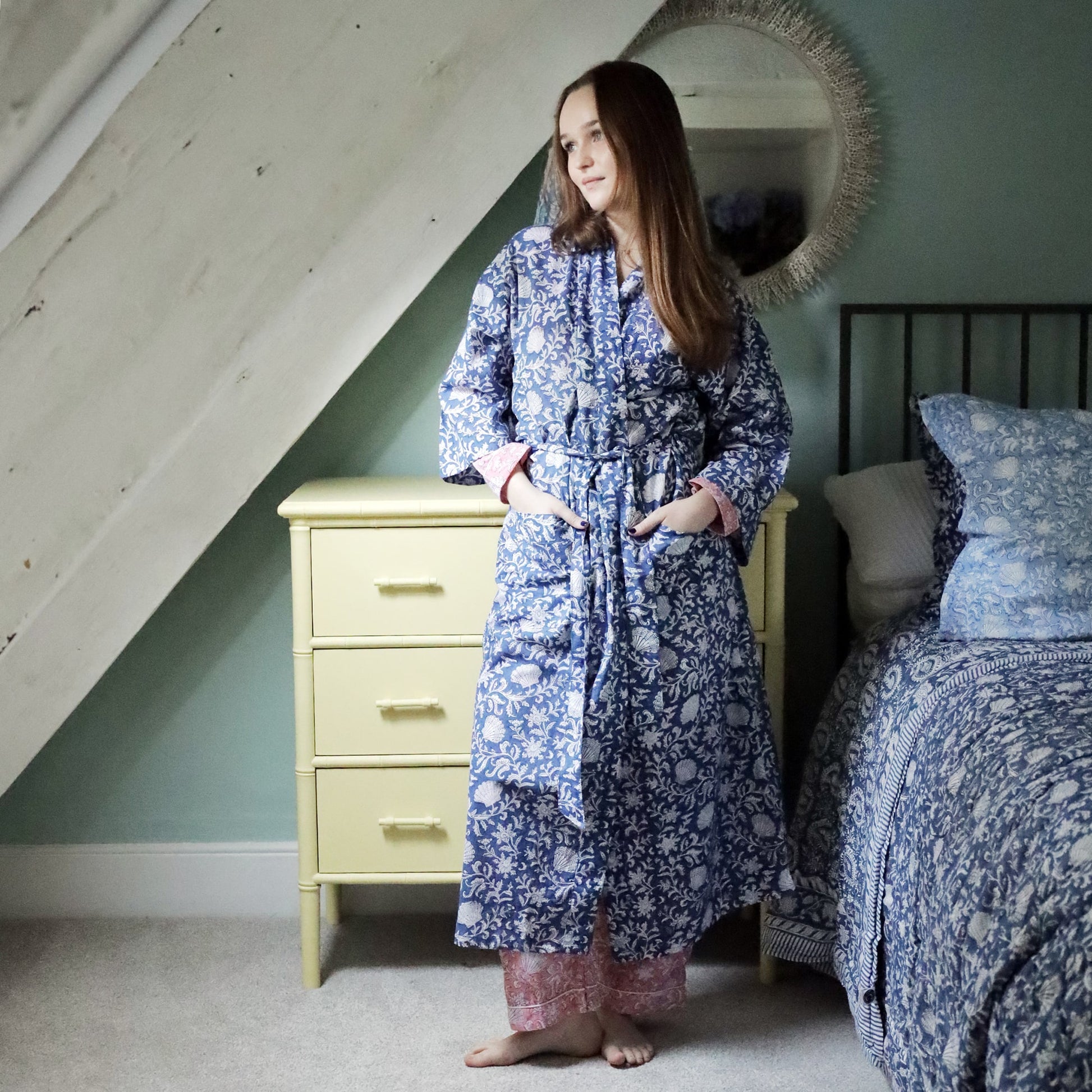 A woman wearing a navy blue dressing gown with intricate white block-printed floral and shell patterns, tied at the waist with a matching belt. She stands barefoot in a cozy bedroom with a soft pastel aesthetic, next to a yellow dresser and a neatly made bed adorned with matching blue bedding.