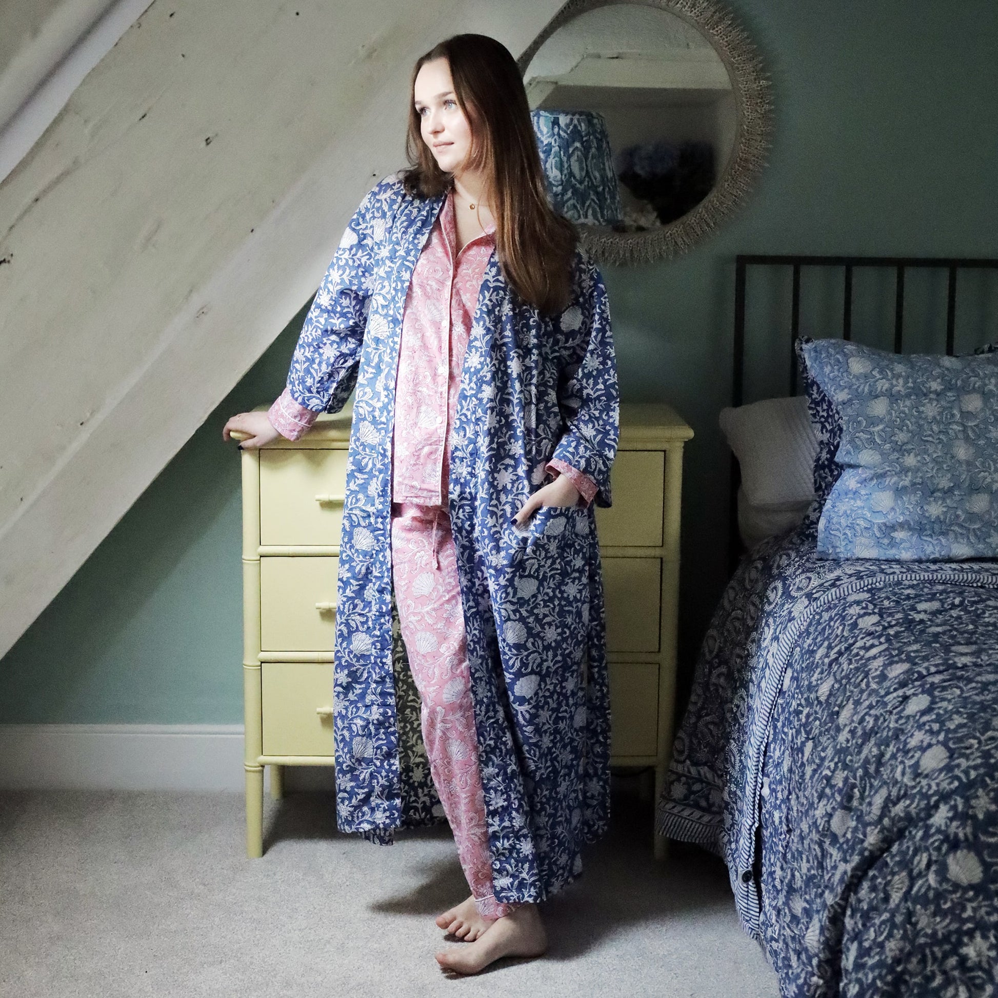 A woman wearing a pink block-printed long dressing gown layered over a navy blue pajama set with white floral patterns. She stands barefoot on a carpeted floor next to a yellow dresser and a bed adorned with matching blue and white bedding. The room has a cozy, rustic charm with a slanted ceiling and a decorative mirror in the background.