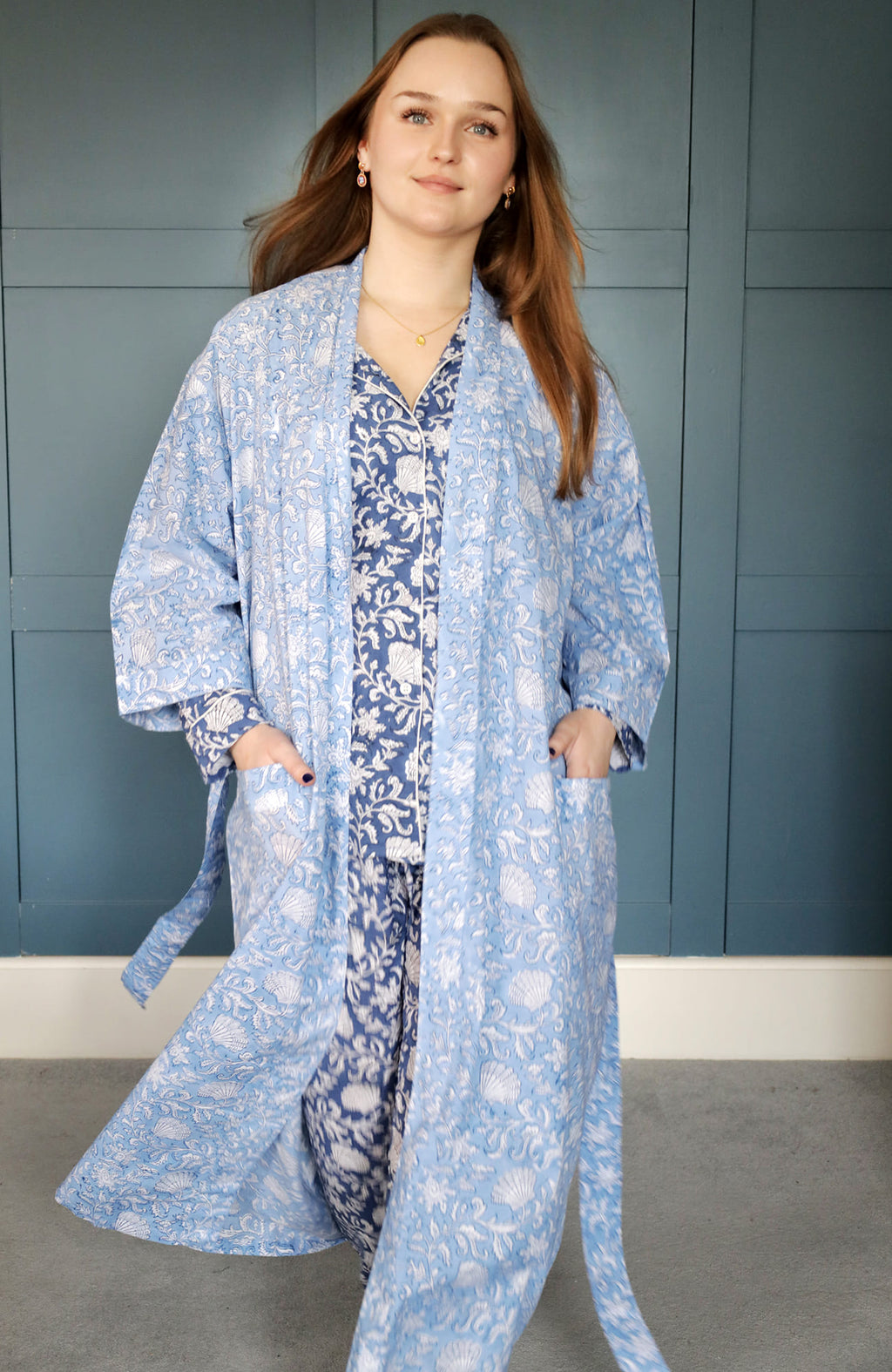 Person posing confidently in a light blue dressing gown featuring intricate white floral and seashell block print patterns. The gown is layered over navy floral pyjamas, set against a teal paneled background for a polished and elegant look.