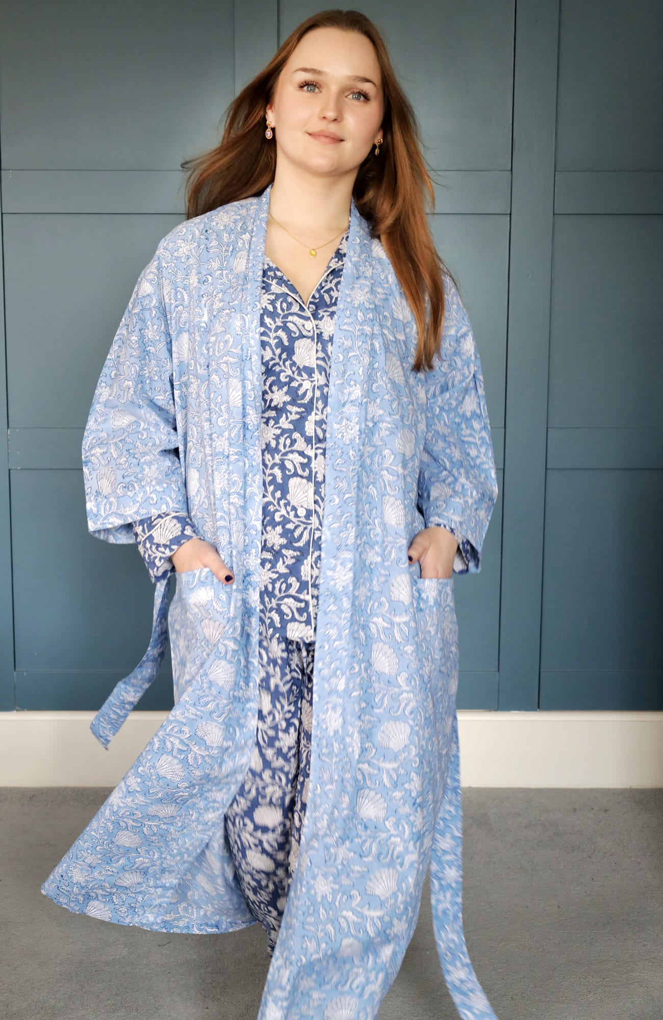 Person posing confidently in a light blue dressing gown featuring intricate white floral and seashell block print patterns. The gown is layered over navy floral pyjamas, set against a teal paneled background for a polished and elegant look.