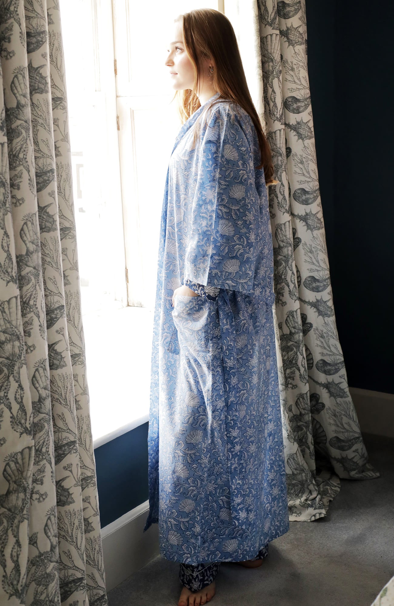 Person standing by a bright window, wearing a light blue dressing gown with intricate white floral and seashell block print patterns, layered over matching pyjamas. The setting features nautical-themed curtains and a deep blue accent wall, creating a serene and coastal-inspired atmosphere.