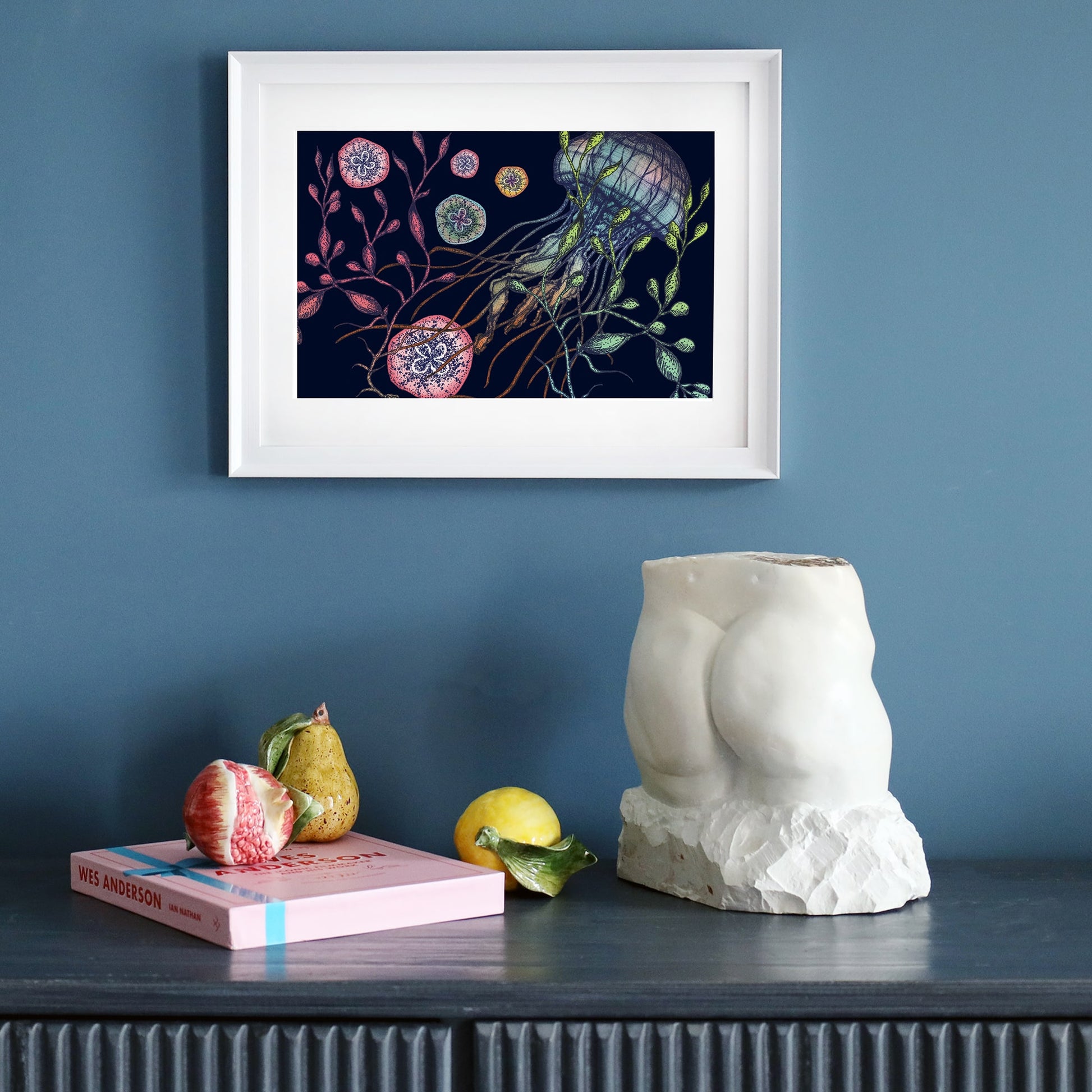 A framed illustration of a jellyfish with seaweed and marine elements on a dark navy background, set in a white frame with a white mount. The artwork is displayed on a dark sideboard, alongside a sculptural torso, ceramic fruit, and a pink book.