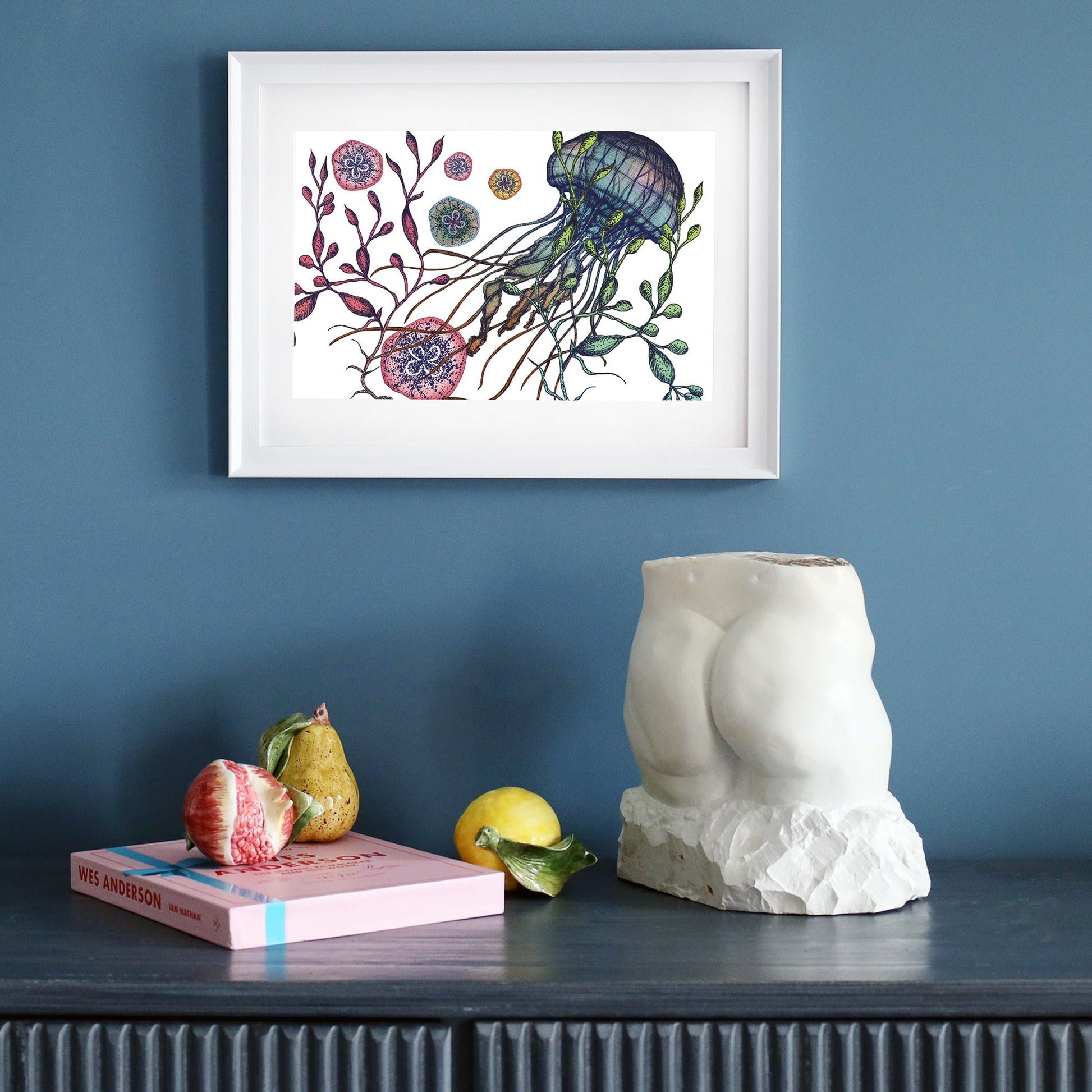 A framed jellyfish illustration hangs on a deep blue wall above a dark sideboard. Below, a sculptural torso, a pink book, ceramic fruit, and a lemon are arranged as decor.