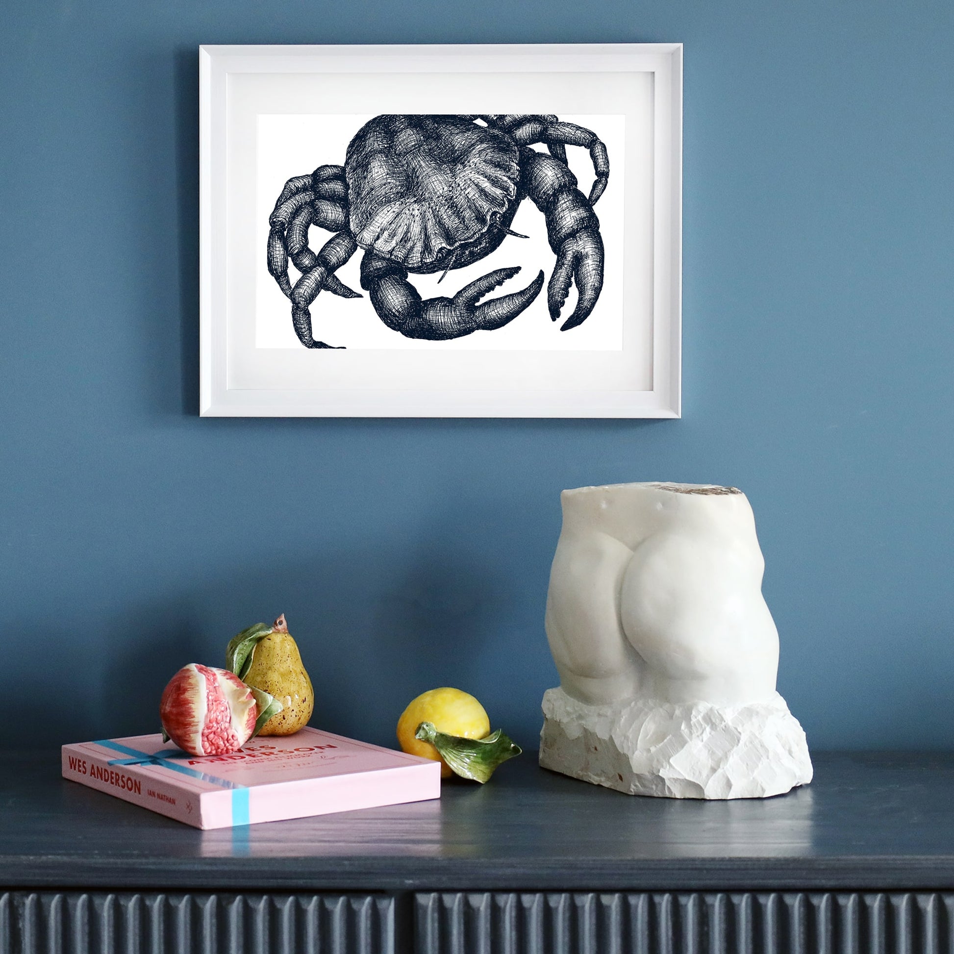 A dark blue wall featuring a framed marine-themed illustration of a crab in a white frame. Below, a black sideboard holds a sculptural white torso, a pink book, ceramic fruit, and a lemon, creating a stylish and artistic display.