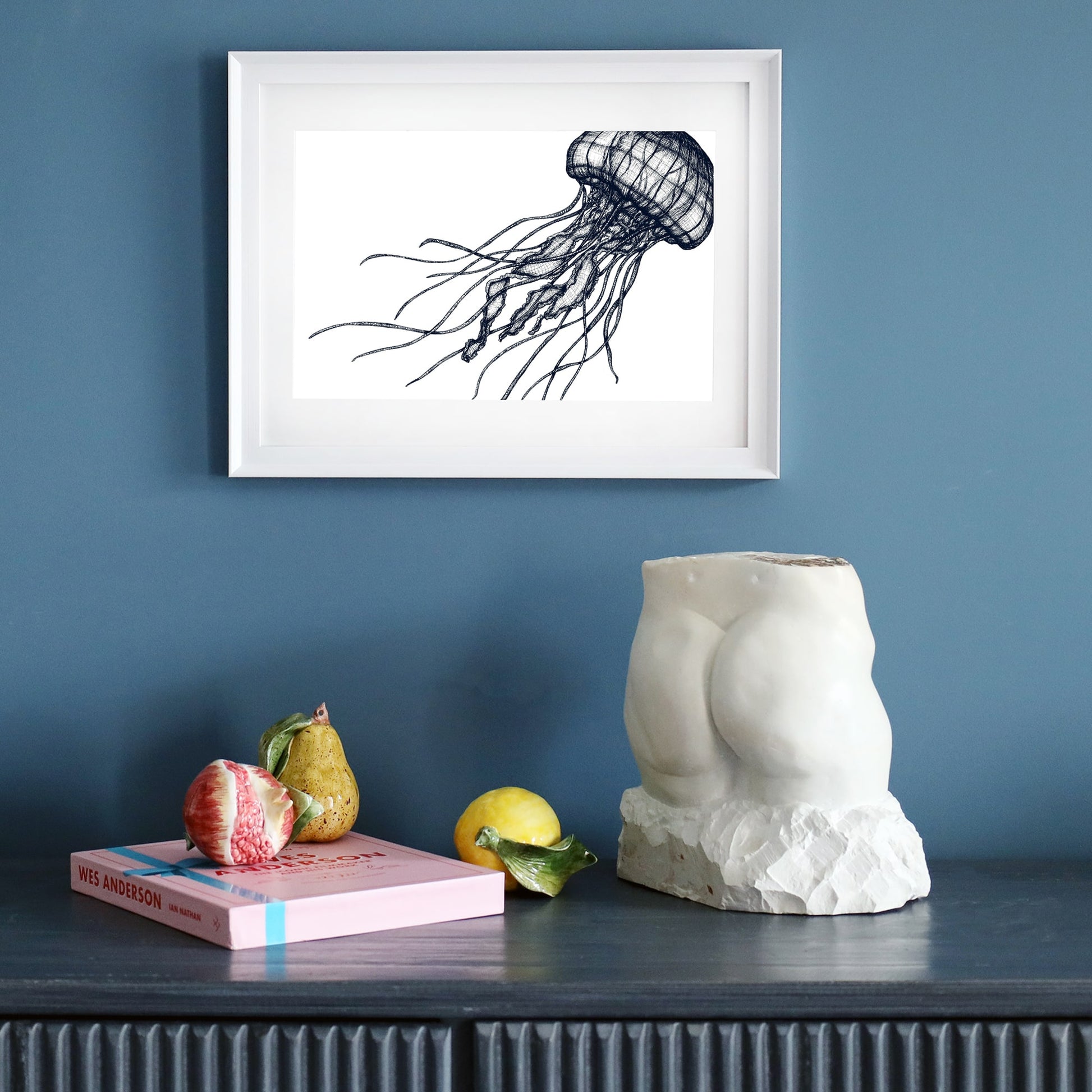 A framed black-and-white jellyfish print on a deep blue wall. Below, a dark wood sideboard holds a white marble torso sculpture, a pink book, ceramic fruit, and a lemon.