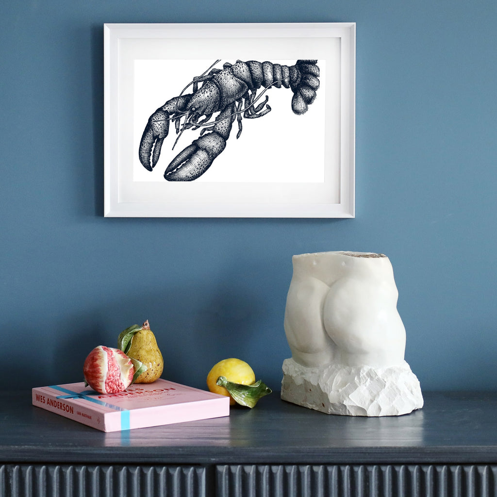 A framed blue-and-white lobster illustration in a white frame, displayed on a dark blue wall. Below, a dark sideboard holds a sculptural torso, a pink book, ceramic fruit, and a yellow lemon.