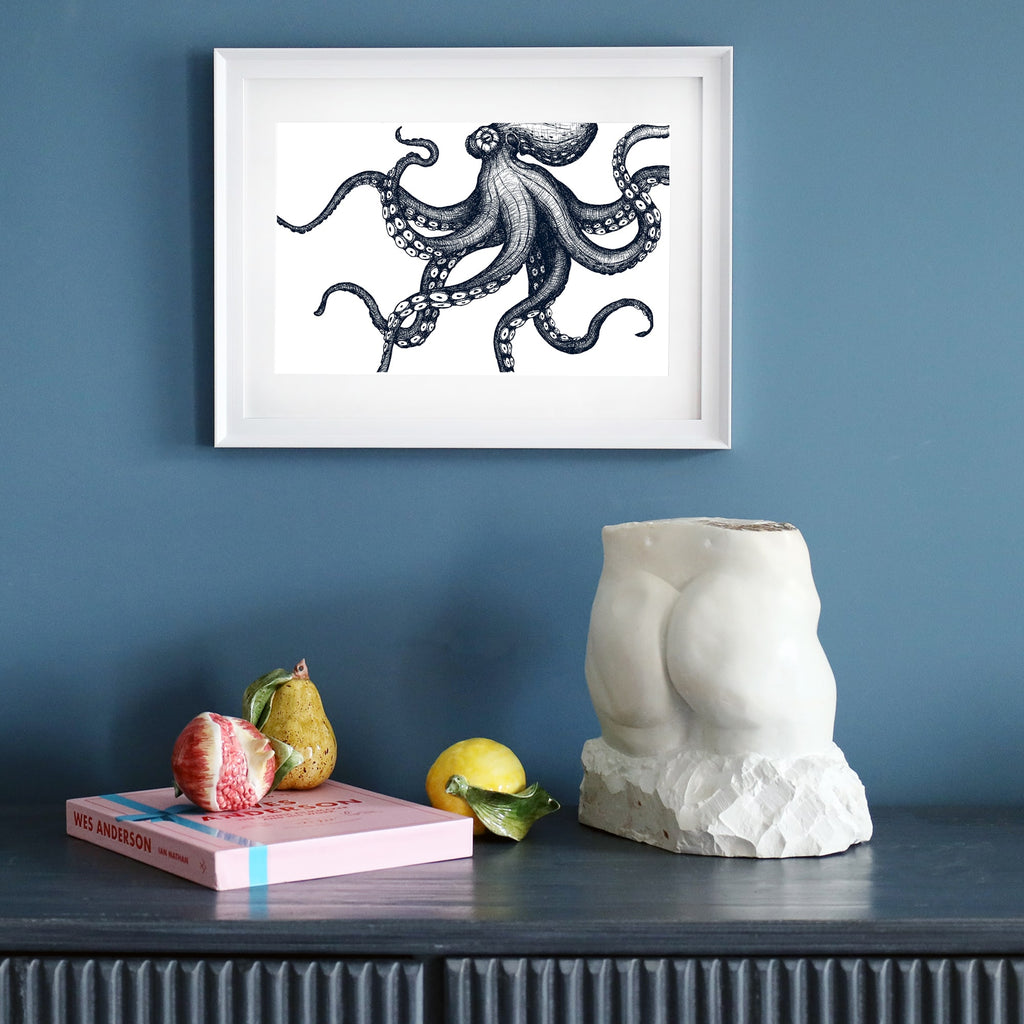 A framed black-and-white octopus illustration in a white frame, displayed on a dark blue wall. Below, a dark sideboard holds a sculptural torso, a pink book, ceramic fruit, and a yellow lemon.