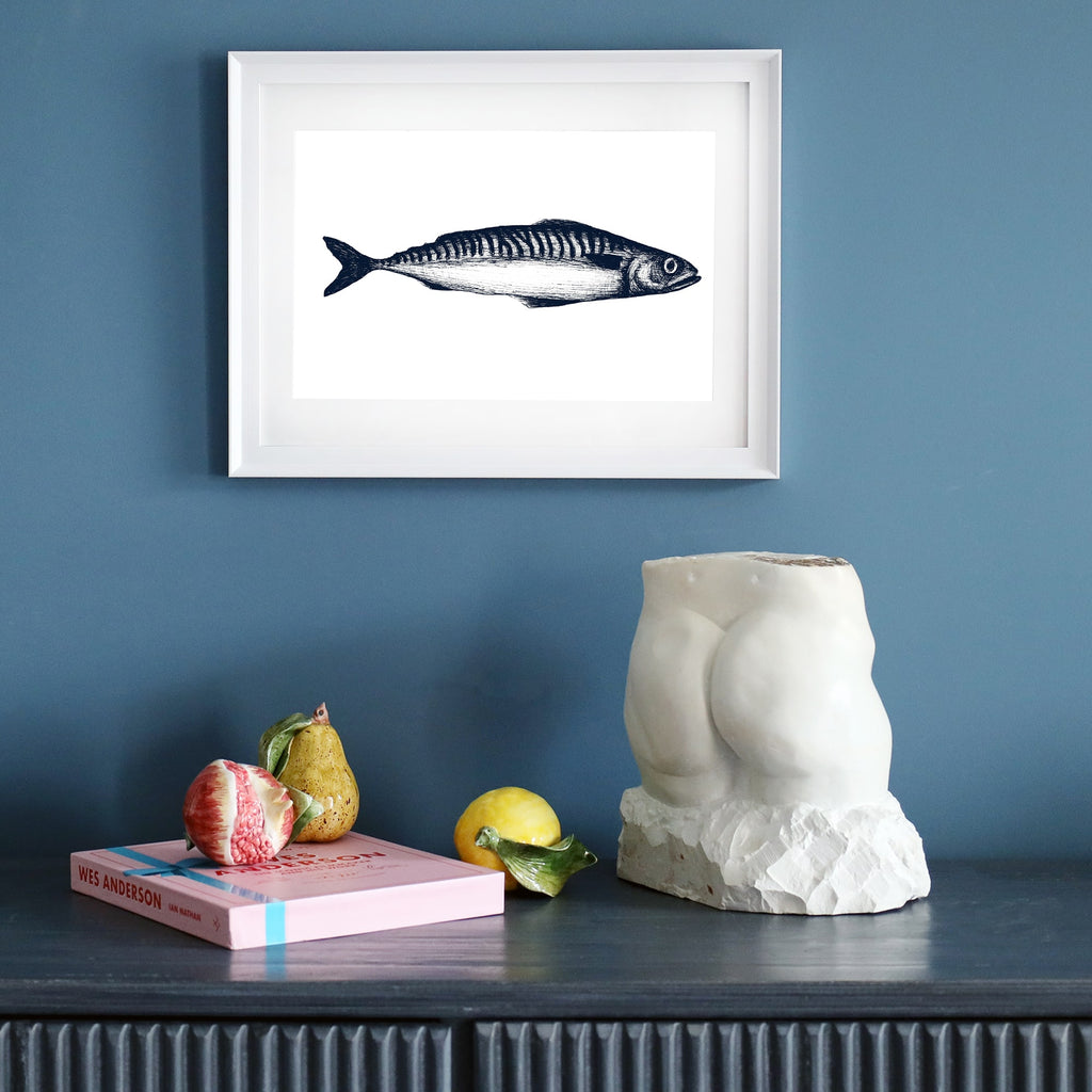 A framed blue-and-white single mackerel illustration in a white frame, displayed on a dark blue wall. Below, a dark sideboard holds a sculptural torso, a pink book, ceramic fruit, and a yellow lemon.