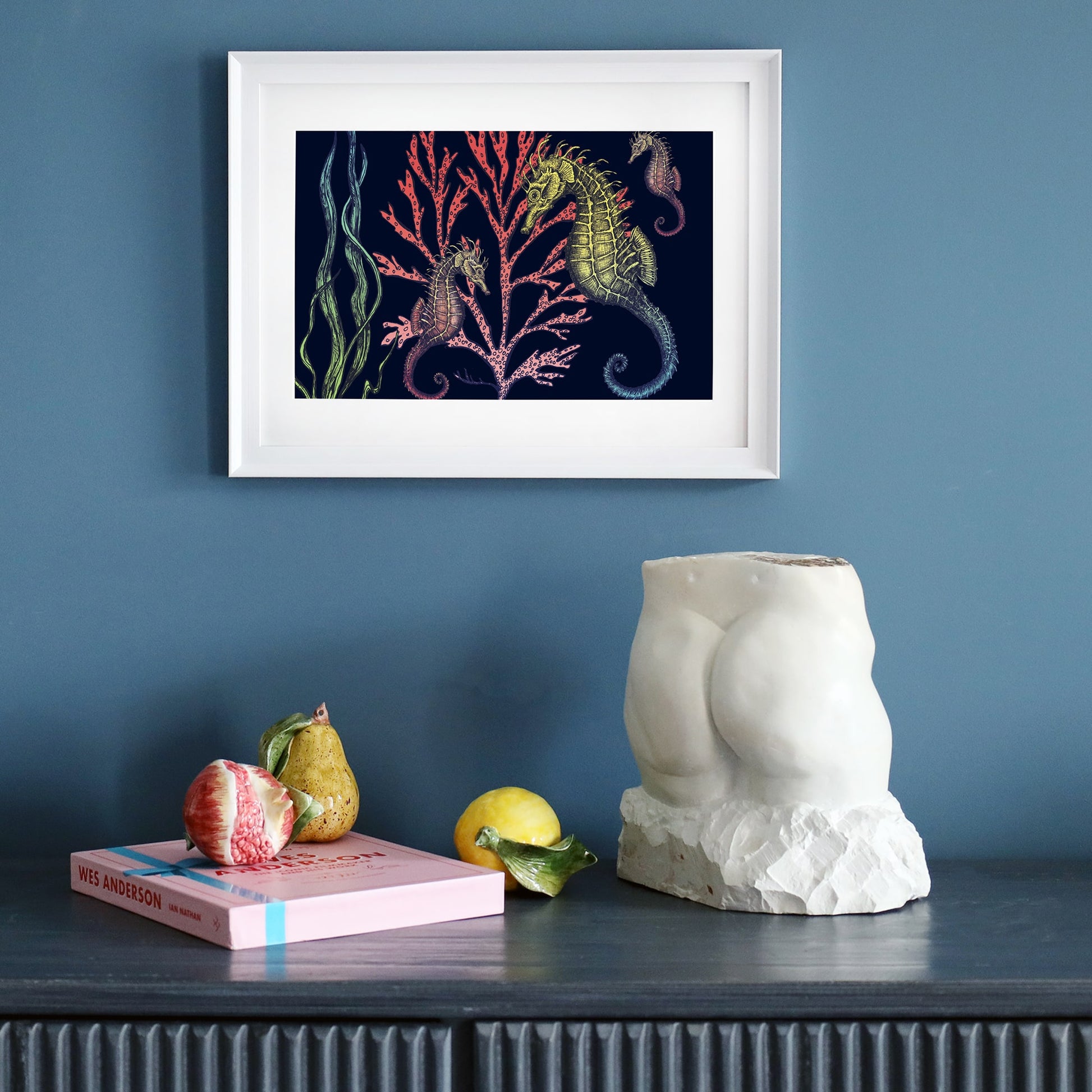 Framed print of seahorses and coral on a navy background, hung on a deep blue wall. Below, a dark wooden sideboard holds a sculptural torso, a pink book, and ceramic fruit.