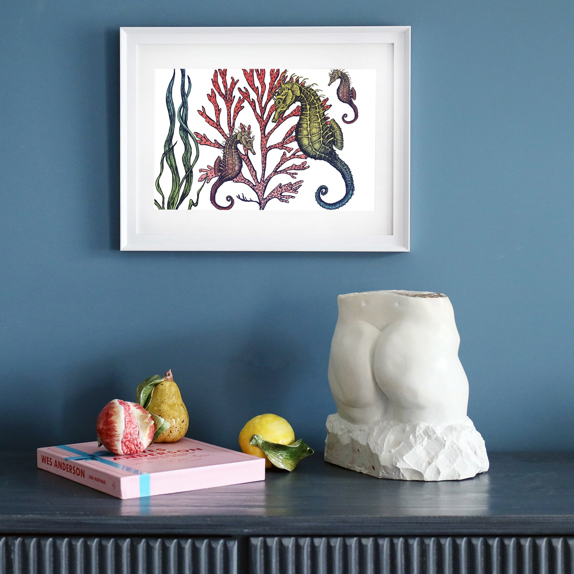  A framed illustration of seahorses among red coral and green seaweed, mounted on a deep blue wall. Below, a dark sideboard holds a white sculpture, a pink book, and ceramic fruits including a fig, pear, and lemon.