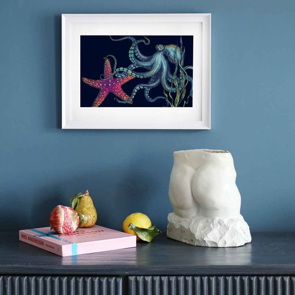 A framed illustration of an octopus and a vibrant red starfish set against a deep navy background hangs on a dark blue wall. The octopus has flowing tentacles detailed in shades of blue and green, surrounded by swaying seaweed. Below the artwork, a dark wooden sideboard holds a sculptural torso, a pink book, ceramic fruits, and a lemon. The minimalist white frame contrasts with the dark background of the print.