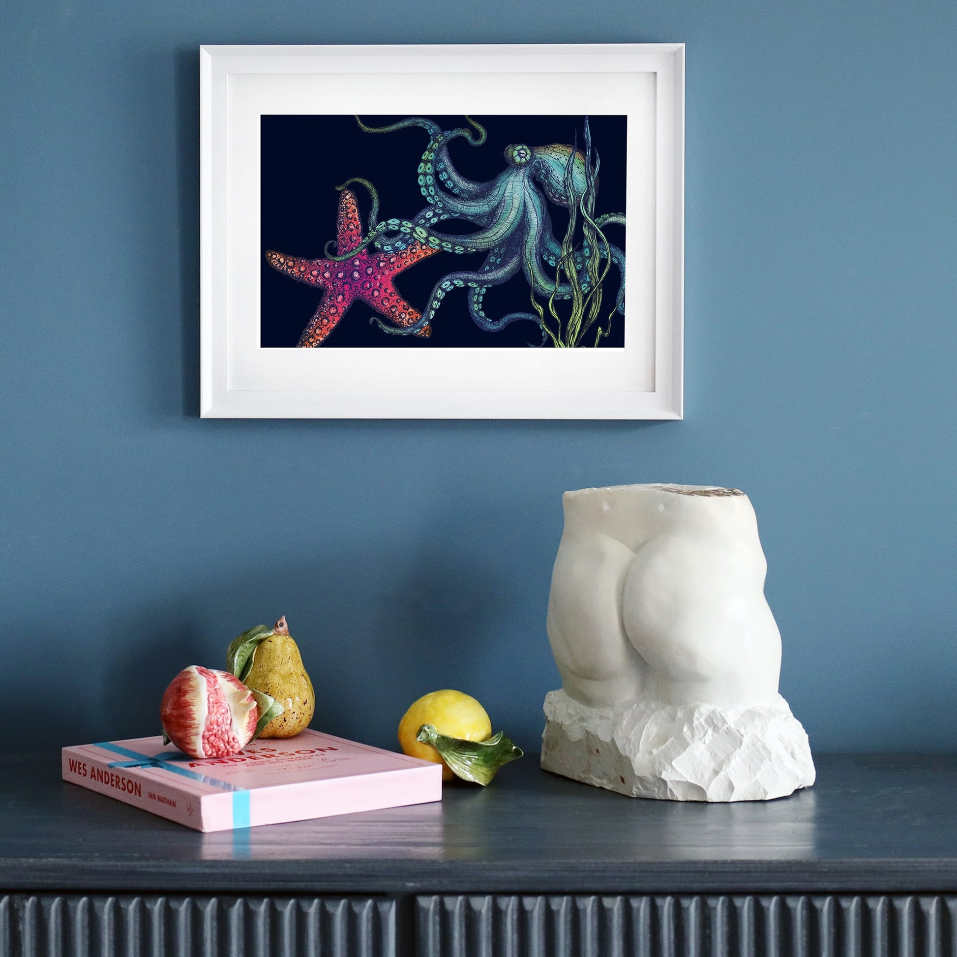 A framed illustration of an octopus and a vibrant red starfish set against a deep navy background hangs on a dark blue wall. The octopus has flowing tentacles detailed in shades of blue and green, surrounded by swaying seaweed. Below the artwork, a dark wooden sideboard holds a sculptural torso, a pink book, ceramic fruits, and a lemon. The minimalist white frame contrasts with the dark background of the print.