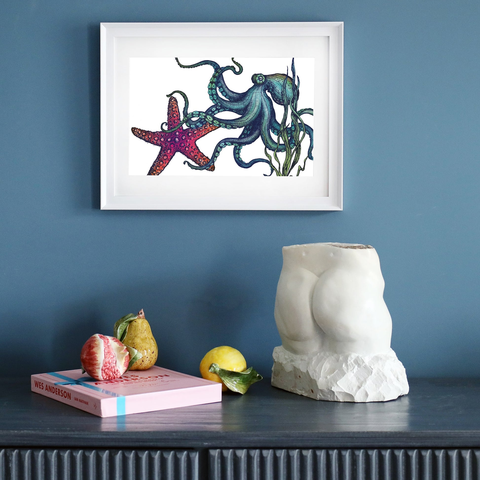A dark blue wall featuring a framed marine-themed illustration of an octopus and a starfish in a white frame. Below, a black sideboard holds a sculptural white torso, a pink book, ceramic fruit, and a lemon, creating a stylish and artistic display.