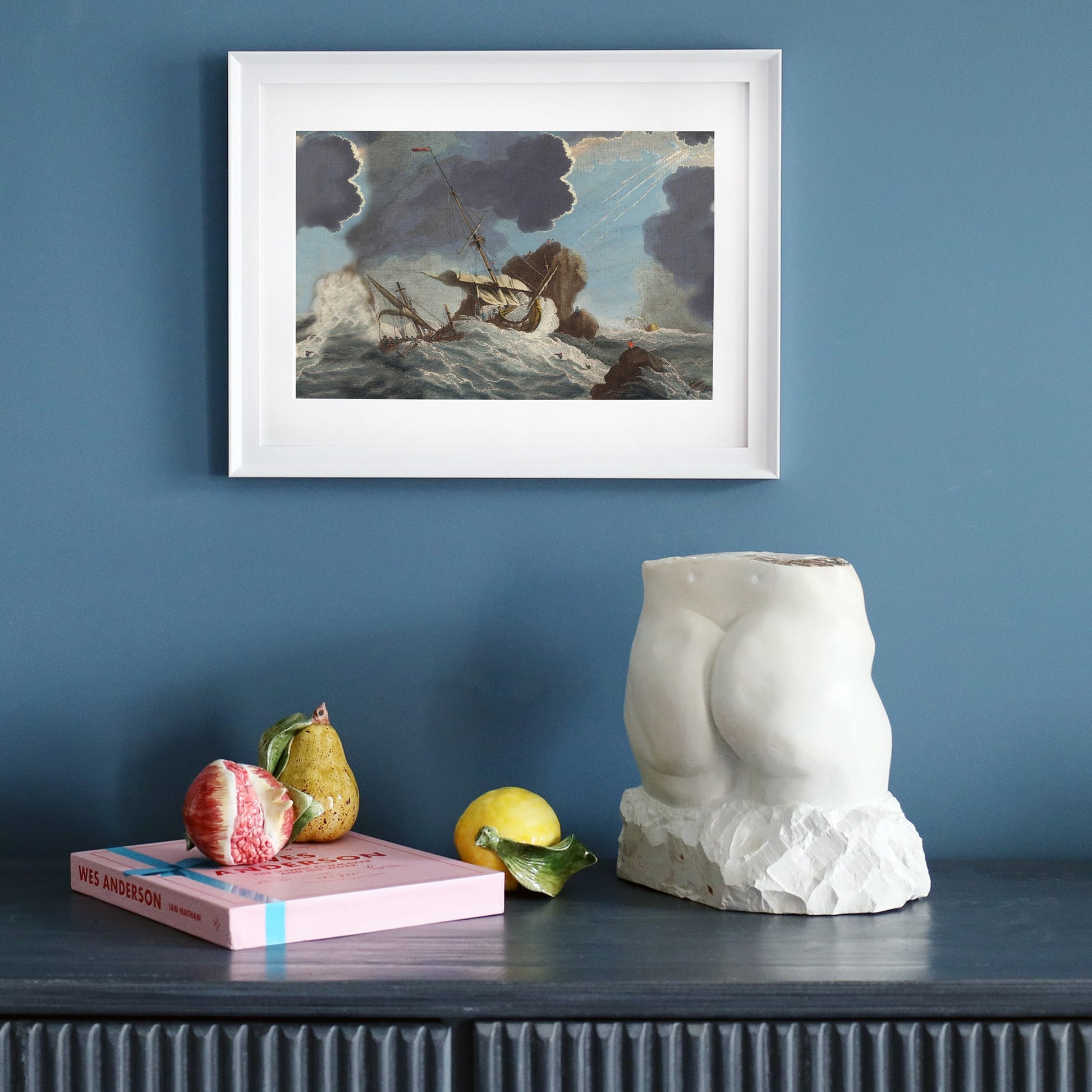 A framed maritime painting of a dramatic shipwreck scene is mounted on a deep blue wall. The artwork, in a white frame, depicts a ship battling towering waves under dark storm clouds. Below, a dark wooden sideboard holds an eclectic arrangement: a classical torso sculpture on a rough stone base, a pastel pink Wes Anderson book, realistic ceramic fruit, and a single lemon leaf. The composition blends vintage nautical aesthetics with modern artistic elements.