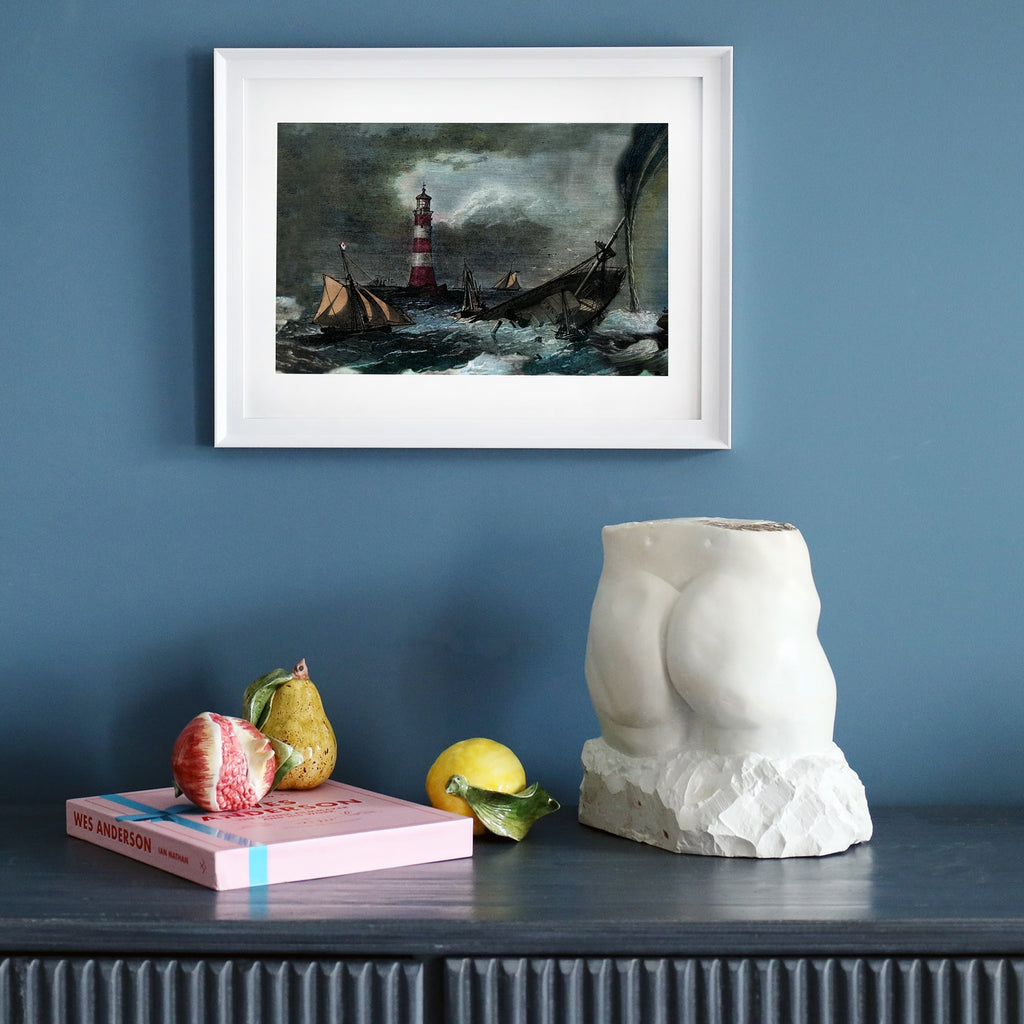 A framed maritime print of a shipwreck near a red-and-white striped lighthouse hangs on a deep blue wall. Below, a dark wooden sideboard holds a sculptural white torso, a pink Wes Anderson book, a pear, a fig, a lemon, and a ceramic leaf, creating an eclectic and artistic display.
