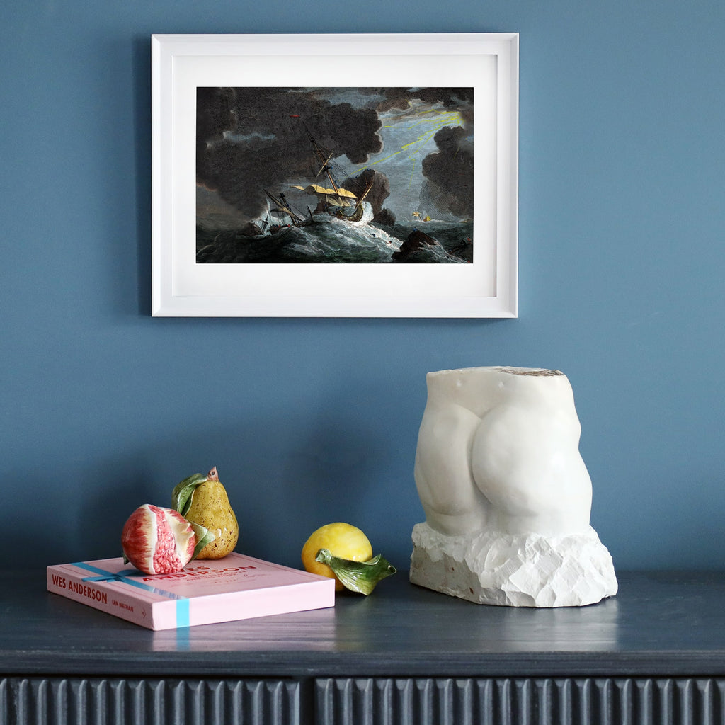Framed print of a stormy shipwreck scene on a dark blue wall, styled on a sideboard with a sculptural torso, decorative fruits, and books.