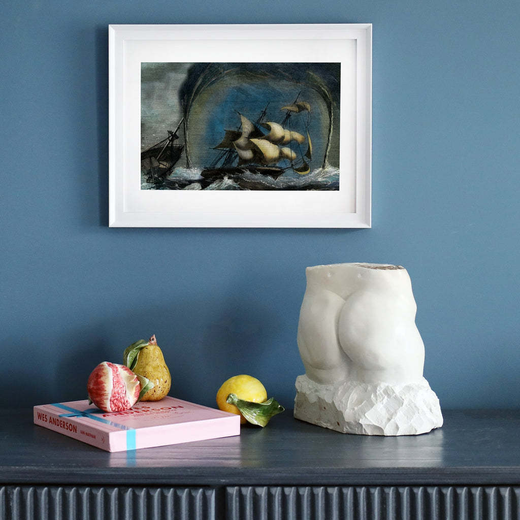 A framed maritime print depicting a dramatic shipwreck amidst a storm and towering waterspouts, displayed on a dark wooden sideboard against a deep blue wall. Below the artwork, a sculptural white torso, a pink Wes Anderson book, and realistic ceramic fruit create an eclectic, artistic arrangement.
