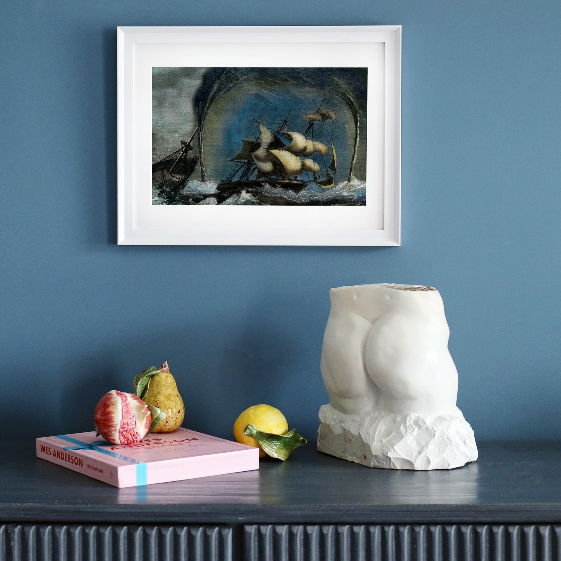 A framed maritime print depicting a dramatic shipwreck amidst a storm and towering waterspouts, displayed on a dark wooden sideboard against a deep blue wall. Below the artwork, a sculptural white torso, a pink Wes Anderson book, and realistic ceramic fruit create an eclectic, artistic arrangement.