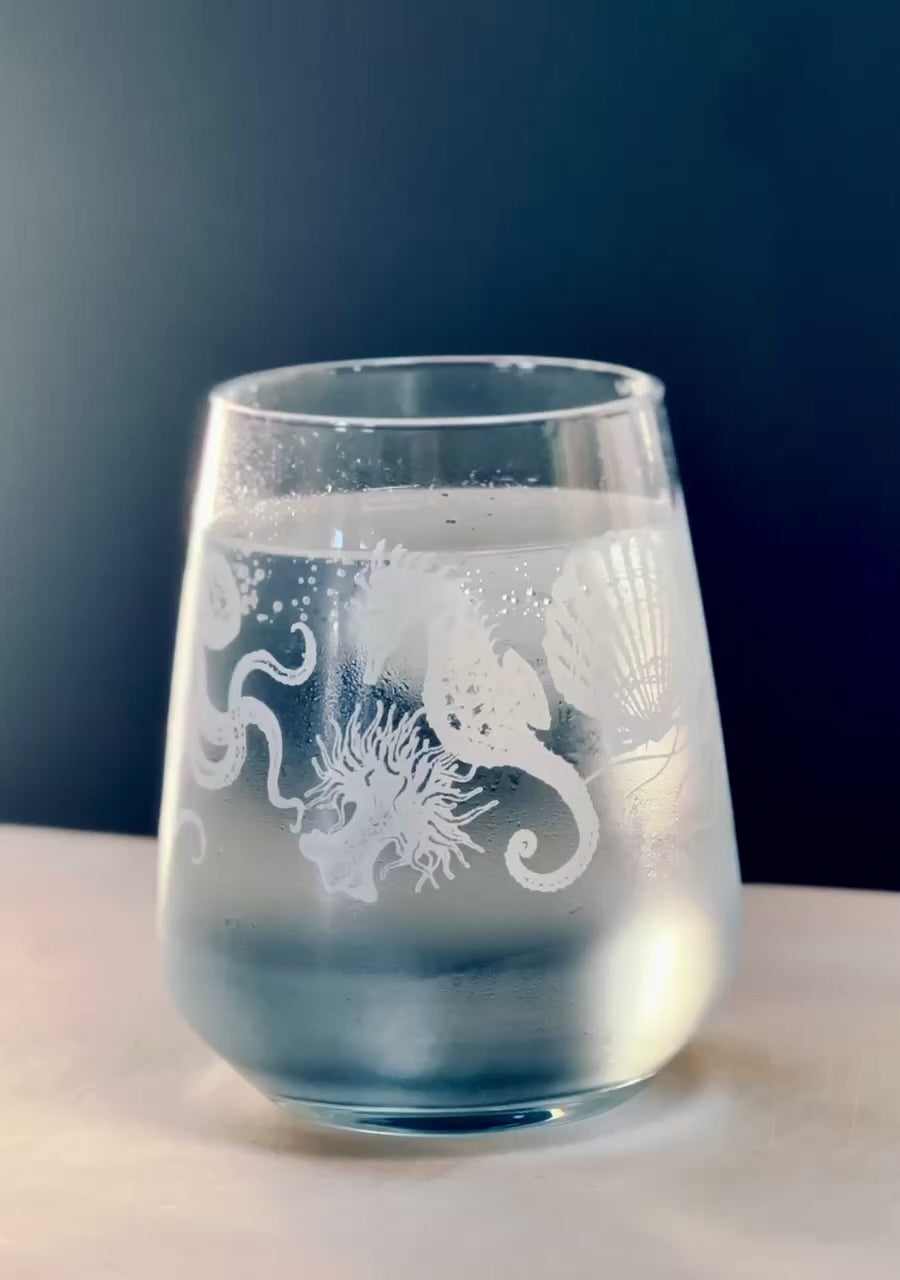 The image shows a clear glass with intricate white prints of underwater sea creatures, including an octopus, jellyfish, seahorse and other marine life. The artwork is detailed and wraps around the glass, creating an aquatic-themed design. There is tonic water being poured into the glass and the bubbles create a fizz around the top.