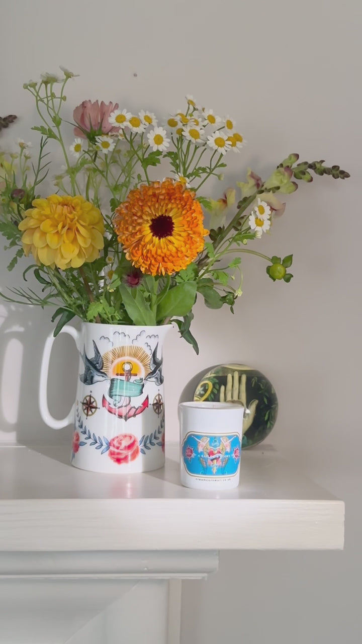 video showing a small candle with mermaid tattoo graphics on a shelf with complimentary jug & flowers and the candle is lit with a match.