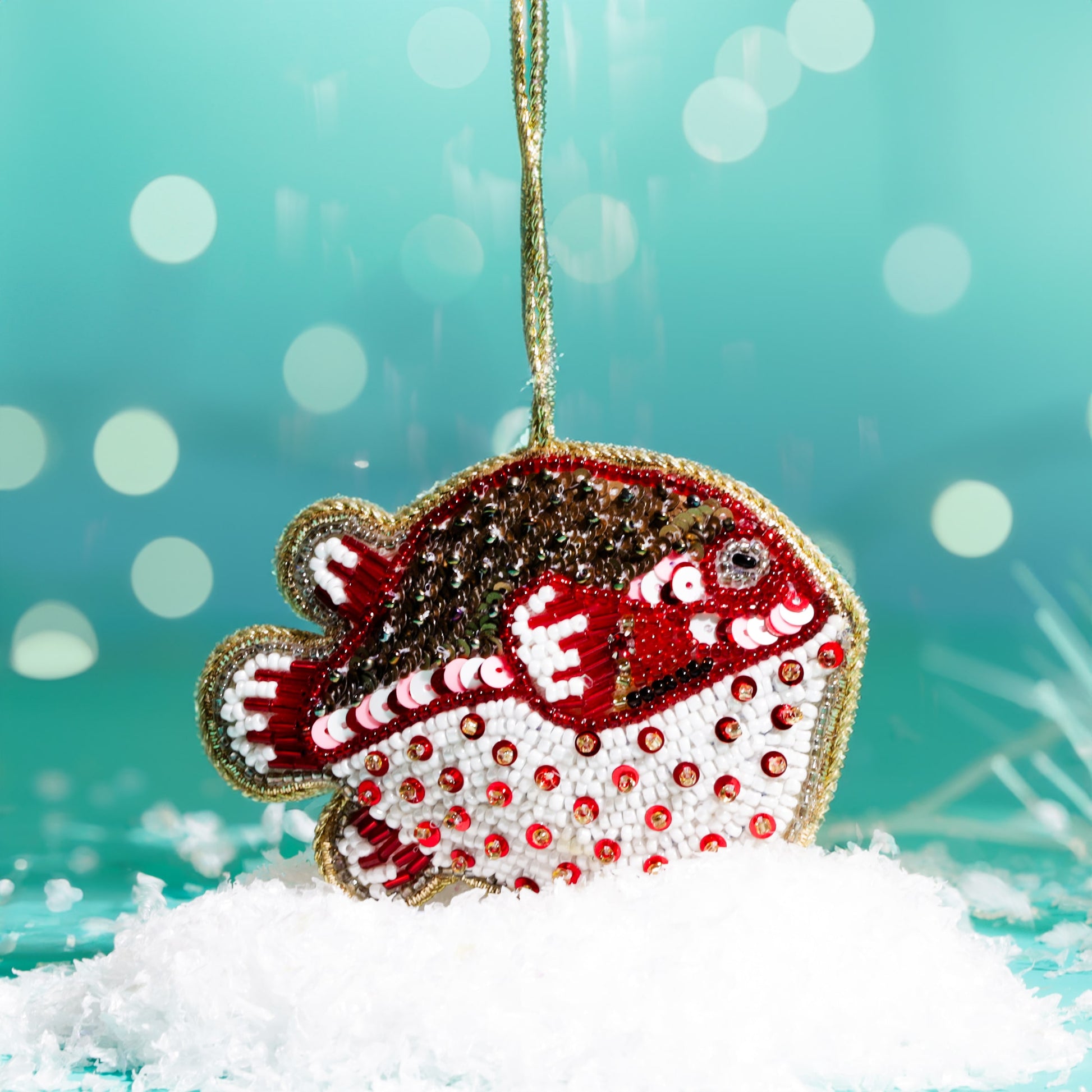 : A red, white, and gold beaded puffer fish decoration with sequins, hanging above artificial snow against a teal background with soft glowing lights.