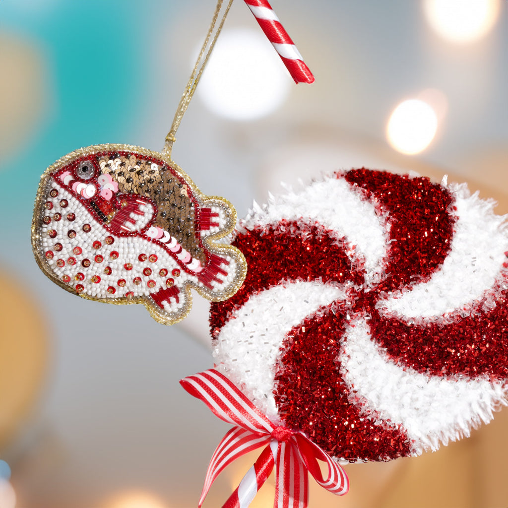 A festive beaded puffer fish decoration in red, white, and gold hanging beside a large red and white glittery lollipop ornament tied with a striped ribbon, against a softly lit background.