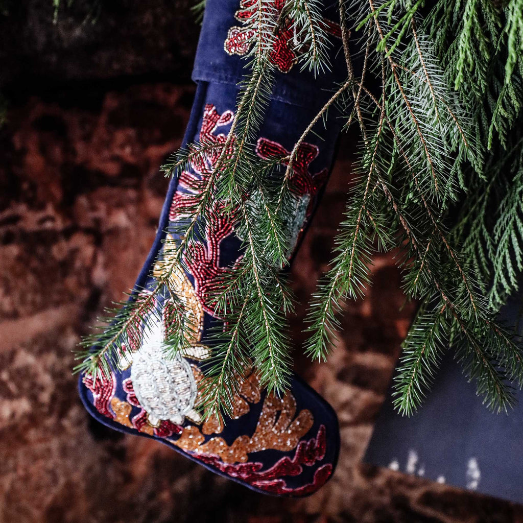 dark blue velvet Christmas stocking with red and orange beaded coral and seaweed and turtle, octopus and lobster floating in amongst it hanging on a foliage covered mantlepiece.