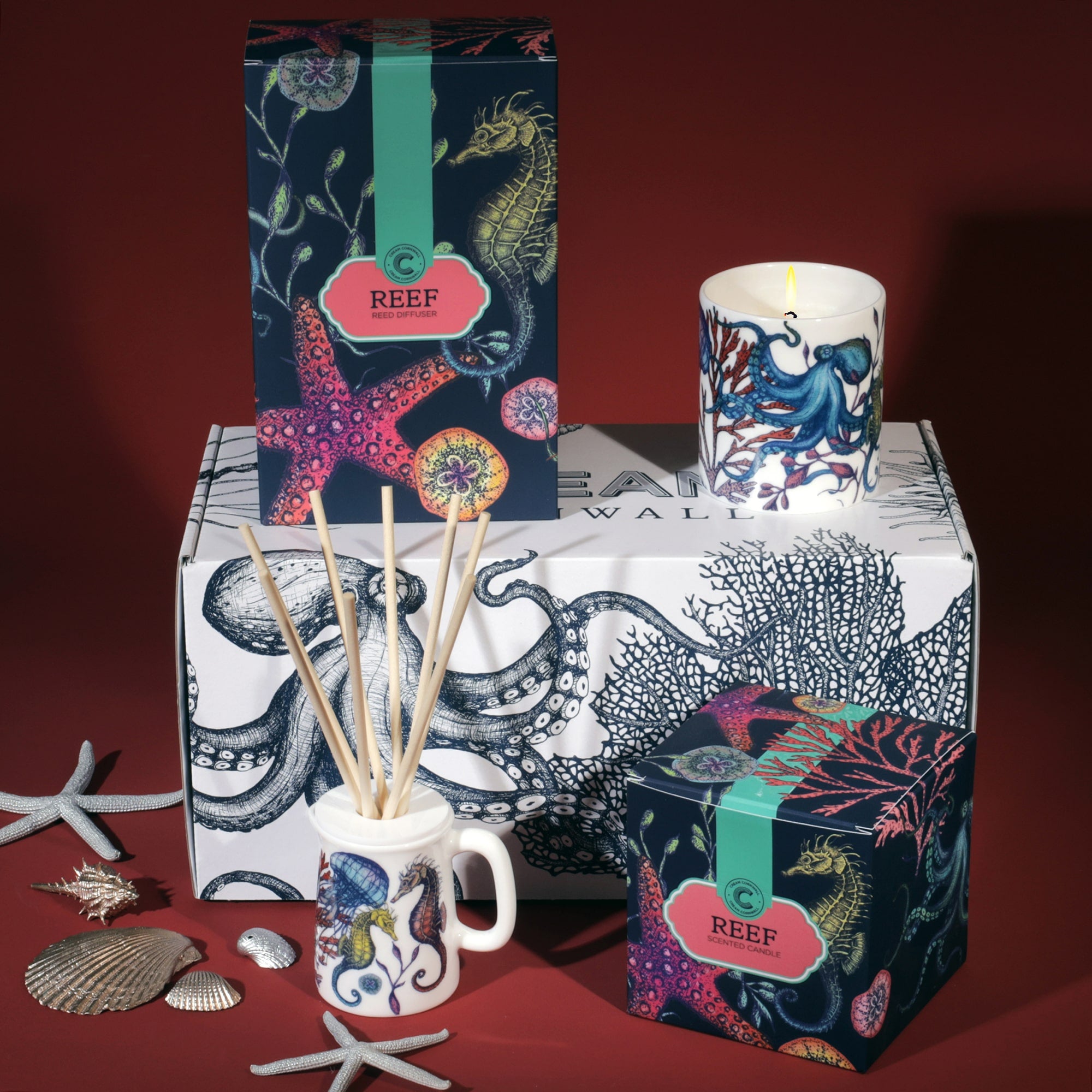 A Cream Cornwall Reef-themed fragrance gift set, featuring a reed diffuser and a scented candle. The packaging and candle holder are decorated with vibrant marine life illustrations, including starfish, seahorses, and octopuses in bright colors. The products are displayed on a box adorned with octopus and coral designs, with decorative seashells and starfish scattered around, enhancing the coastal theme against a deep red background
